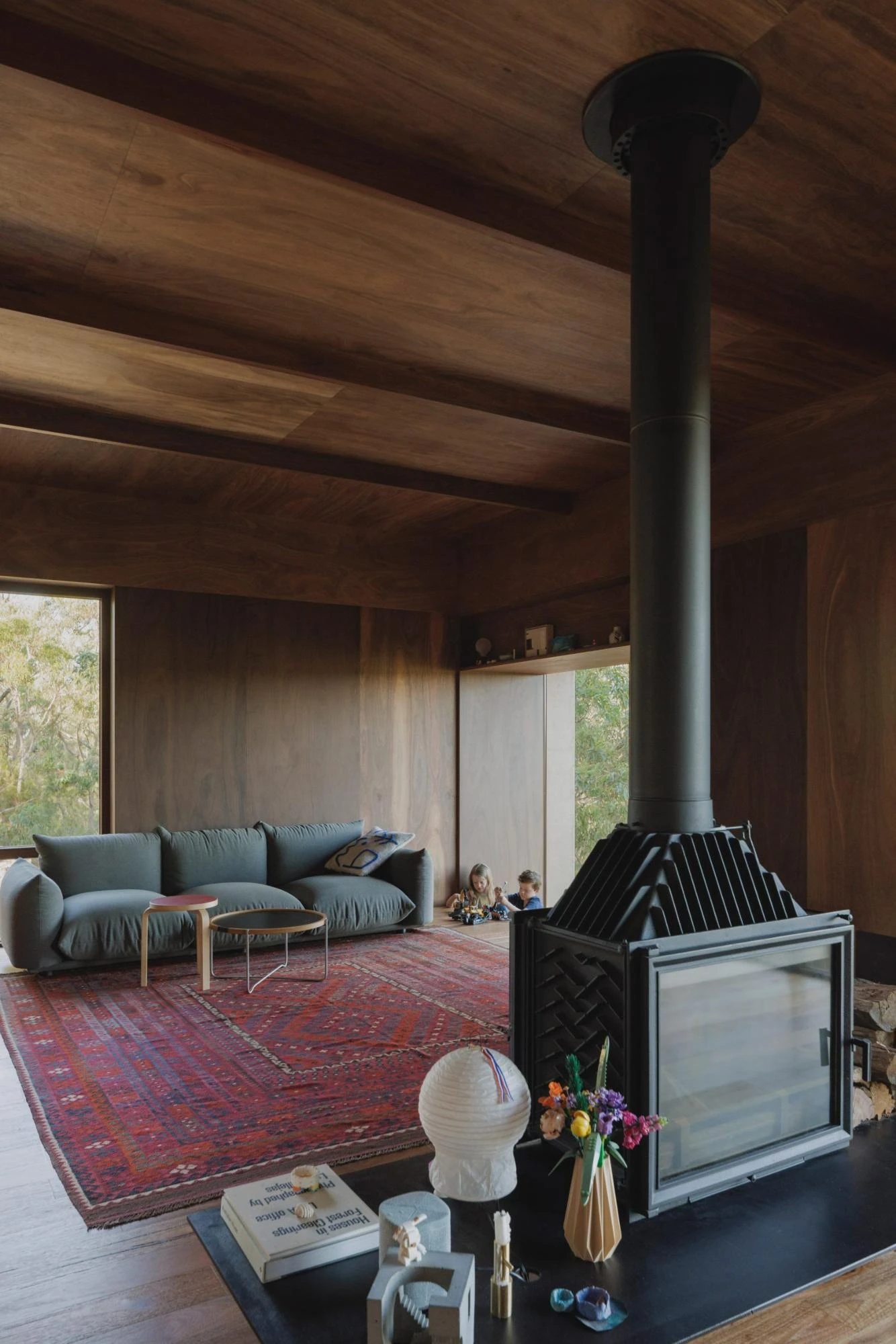 EA House, Southern Highlands by Bokey Grant, photographed by Clinton Weaver, the black firebox and flue rise beside the grey sofa, children reading at a low window, objects gathered on the bench top.