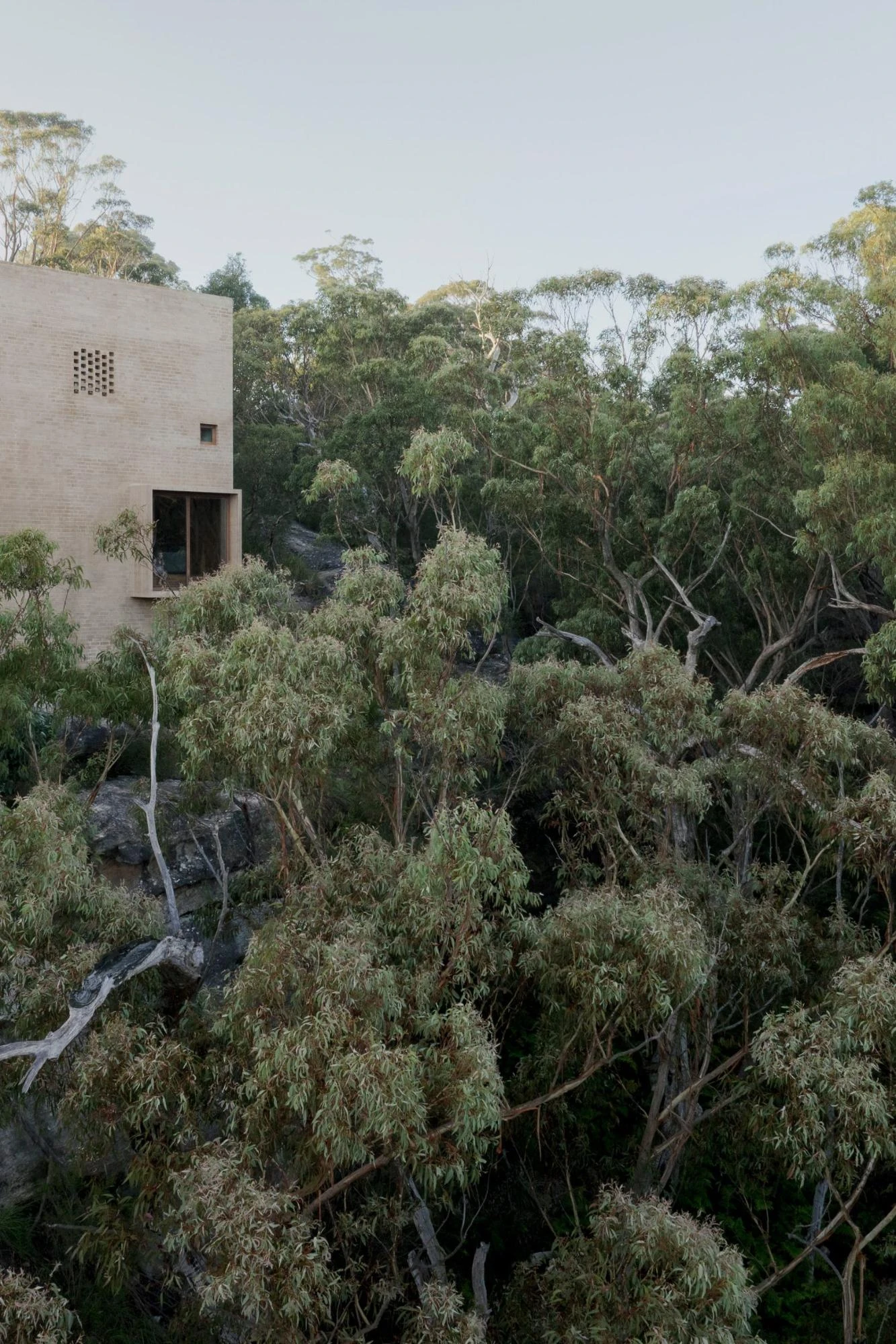 EA House, Southern Highlands by Bokey Grant, photographed by Clinton Weaver, a sand-toned brick volume rises above the eucalypt canopy at dusk, its perforated brick screen catching low light.