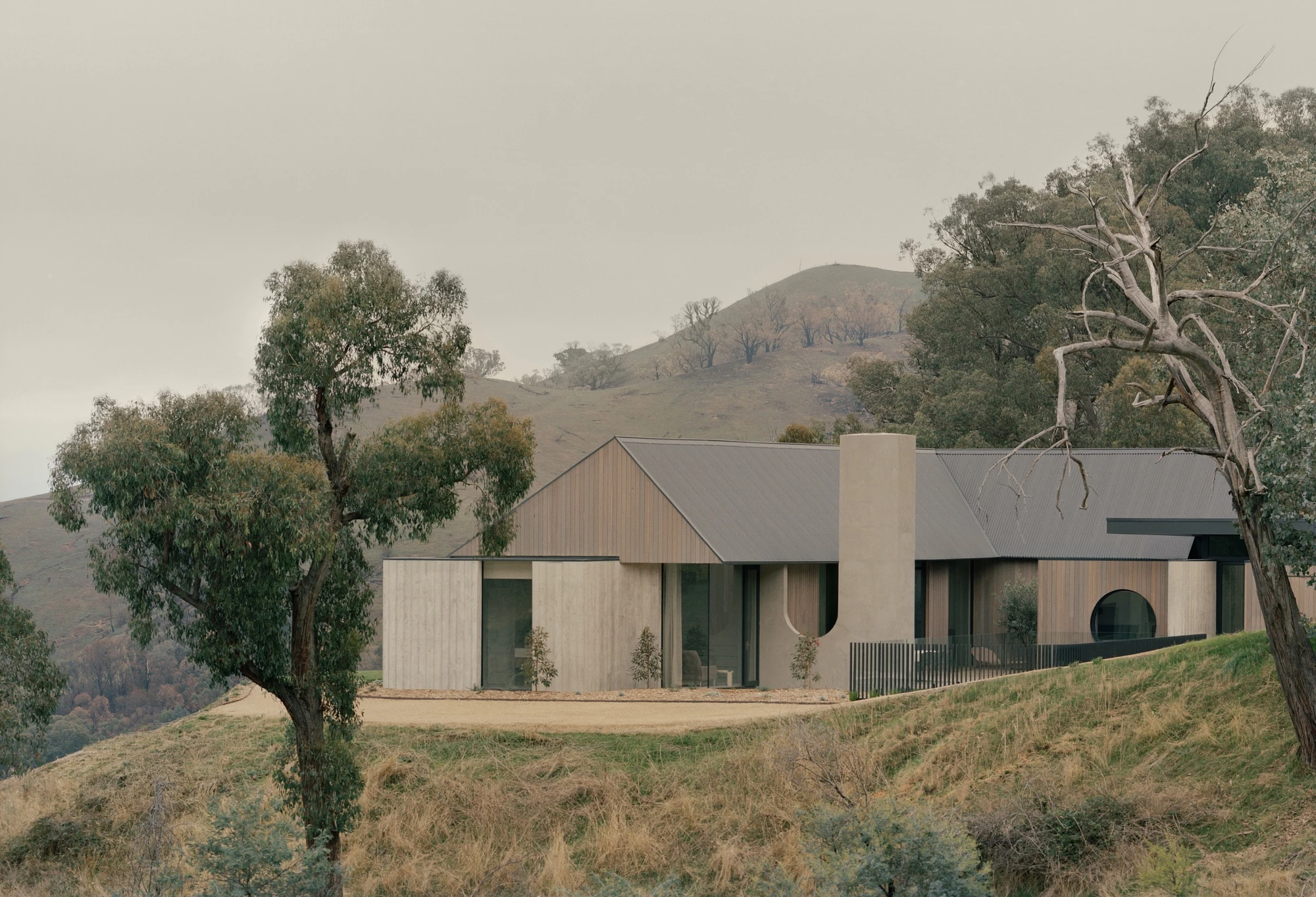 Cathedral House by JAK Architecture, photographed by Tasha Tylee — the house seen from below in overcast light, board-formed concrete and timber cladding under a corrugated gable with a tall plaster chimney and a circular window.