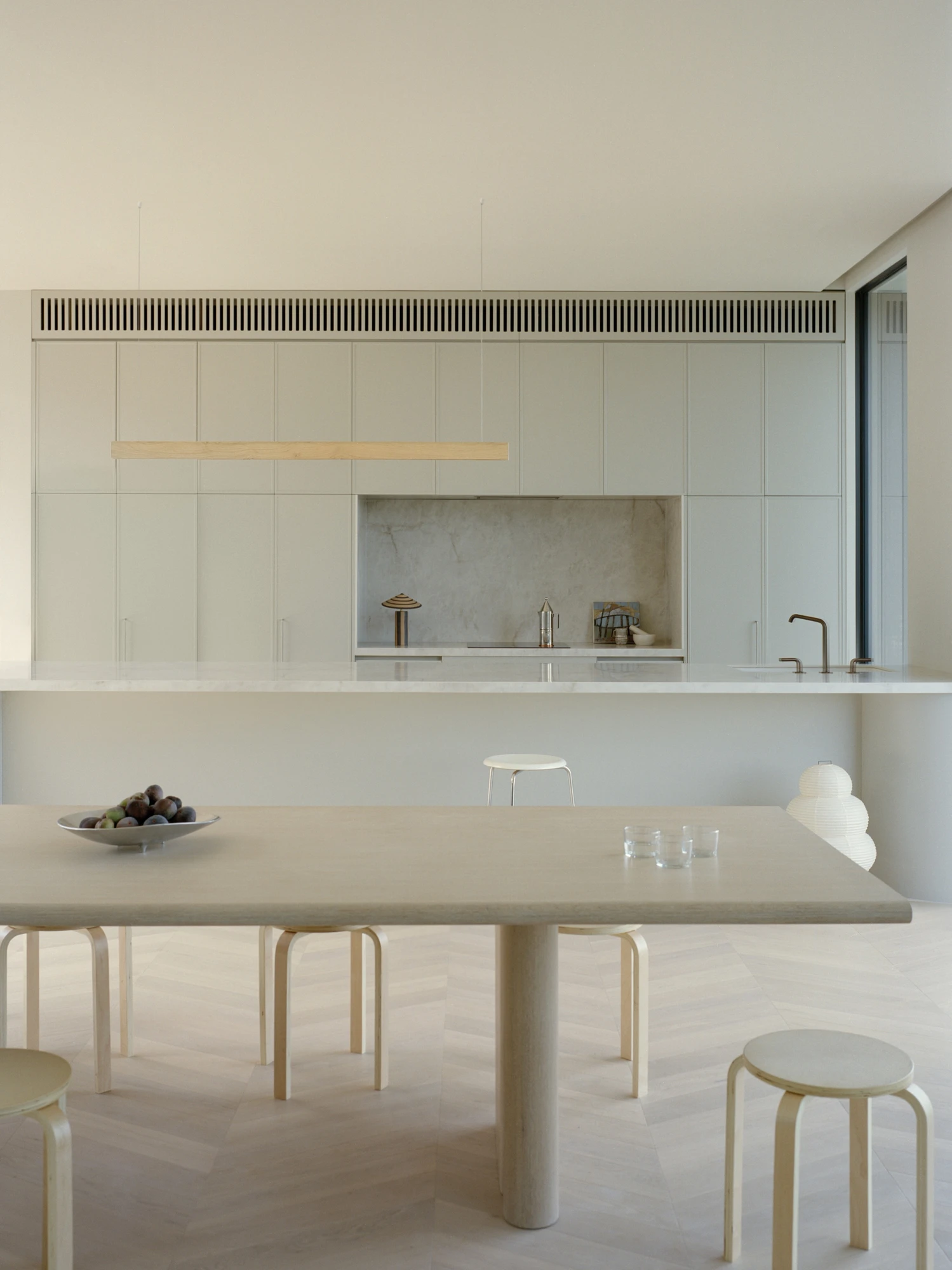 Cathedral House by JAK Architecture, photographed by Tasha Tylee — the kitchen and dining room seen head on, a long pale timber table on a herringbone floor beneath a linear timber pendant, marble island beyond.