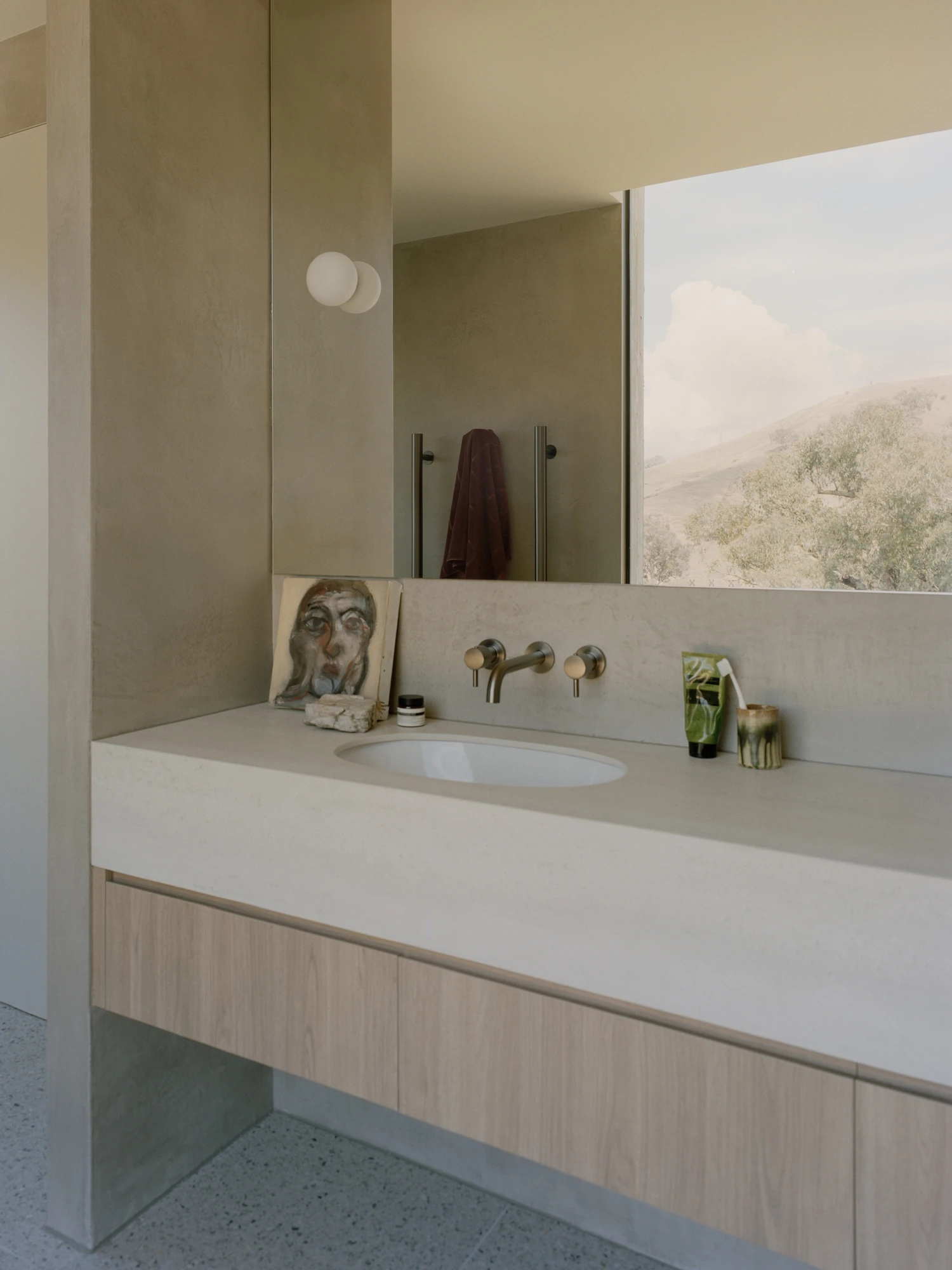 Cathedral House by JAK Architecture, photographed by Tasha Tylee — a plaster ensuite vanity with a stone benchtop and pale timber cabinetry, brushed tapware and a wide mirror reflecting a window onto the hills.