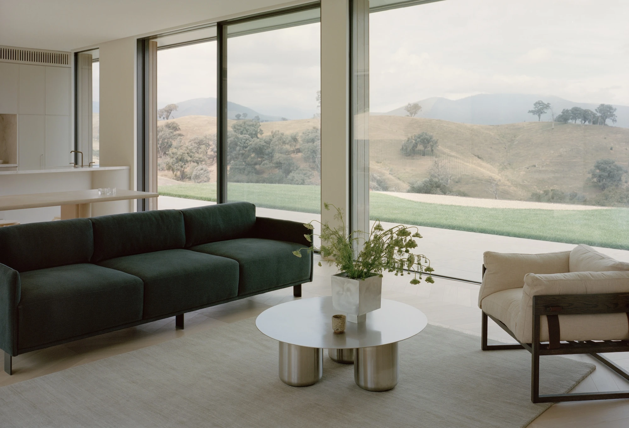 Cathedral House by JAK Architecture, photographed by Tasha Tylee — a living room with a green velvet sofa and pale armchair around a steel coffee table, floor-to-ceiling glazing opening to the ranges.