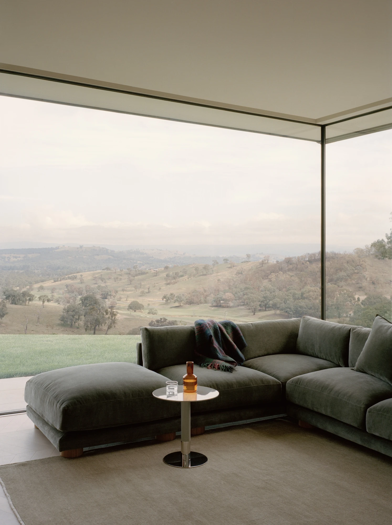 Cathedral House by JAK Architecture, photographed by Tasha Tylee — a green velvet corner sofa on a wool rug set against a frameless glazed corner that opens onto the misty valley and hills.