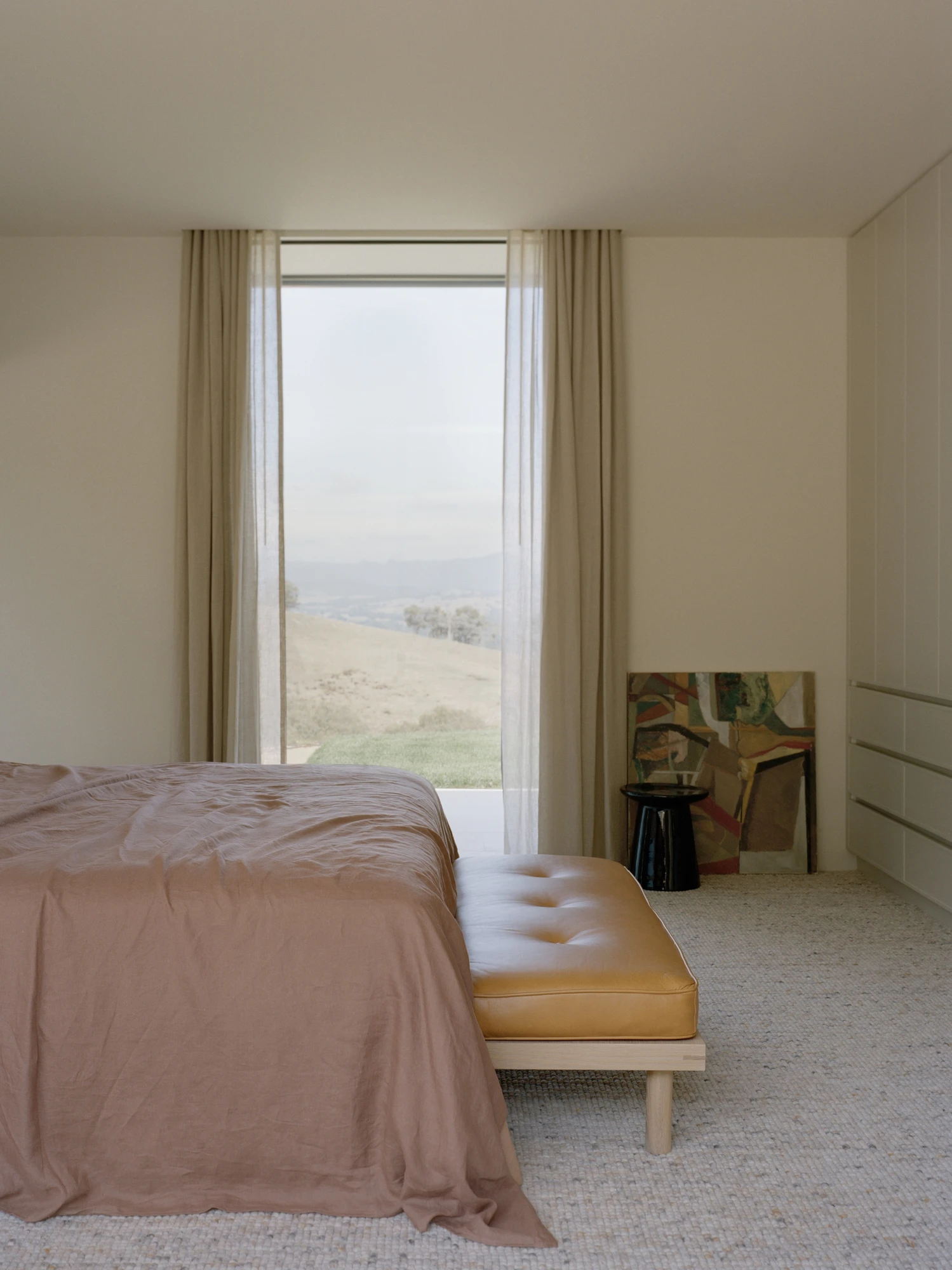 Cathedral House by JAK Architecture, photographed by Tasha Tylee — a bedroom with a dusty pink linen-dressed bed, a tan leather bench, sheer linen curtains at a tall window and an abstract painting leaning on the wall.