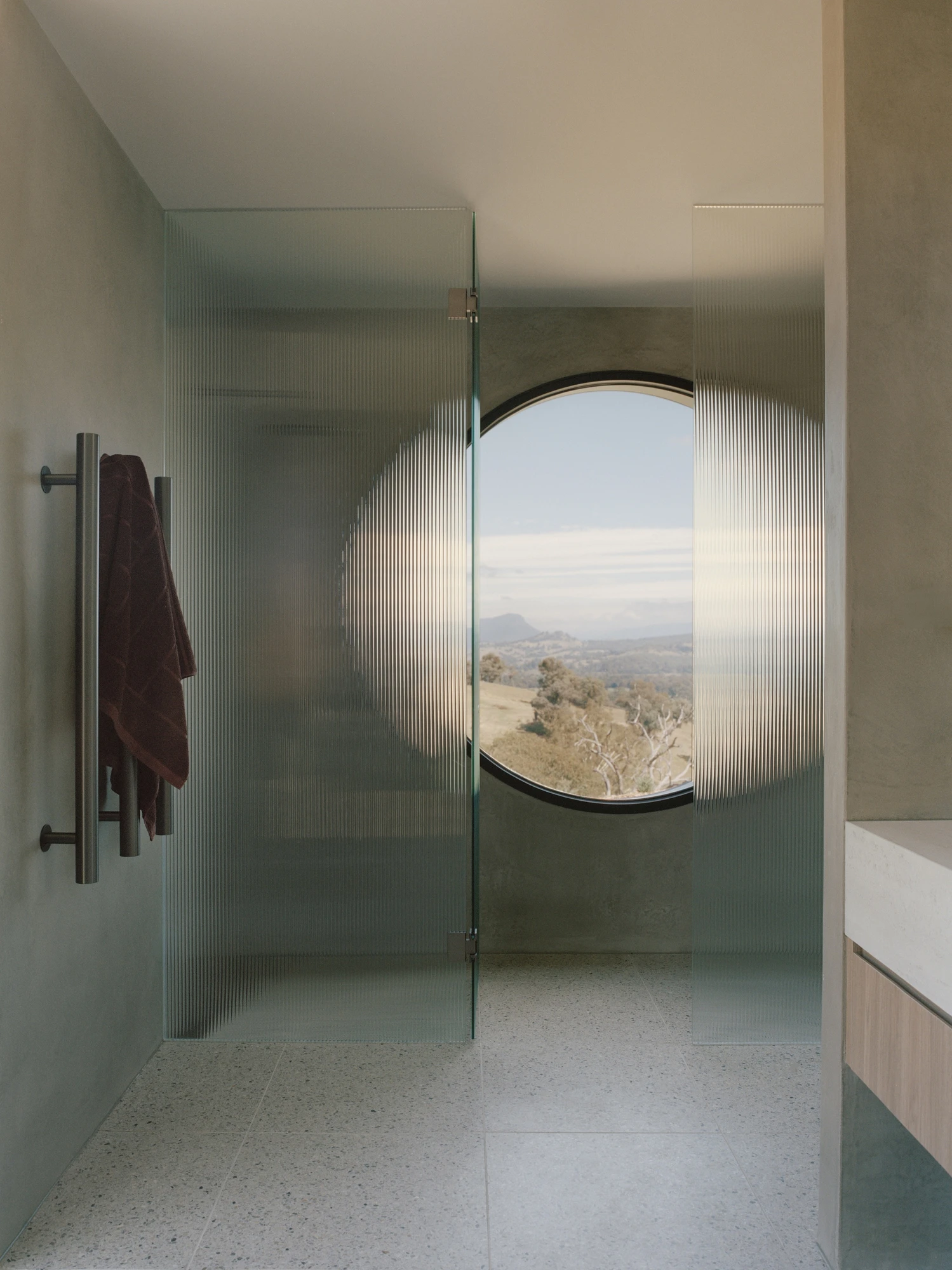 Cathedral House by JAK Architecture, photographed by Tasha Tylee — a terrazzo-floored bathroom where a reeded glass shower screen sits before a circular window framing the ranges, a dark towel hanging on a rail.