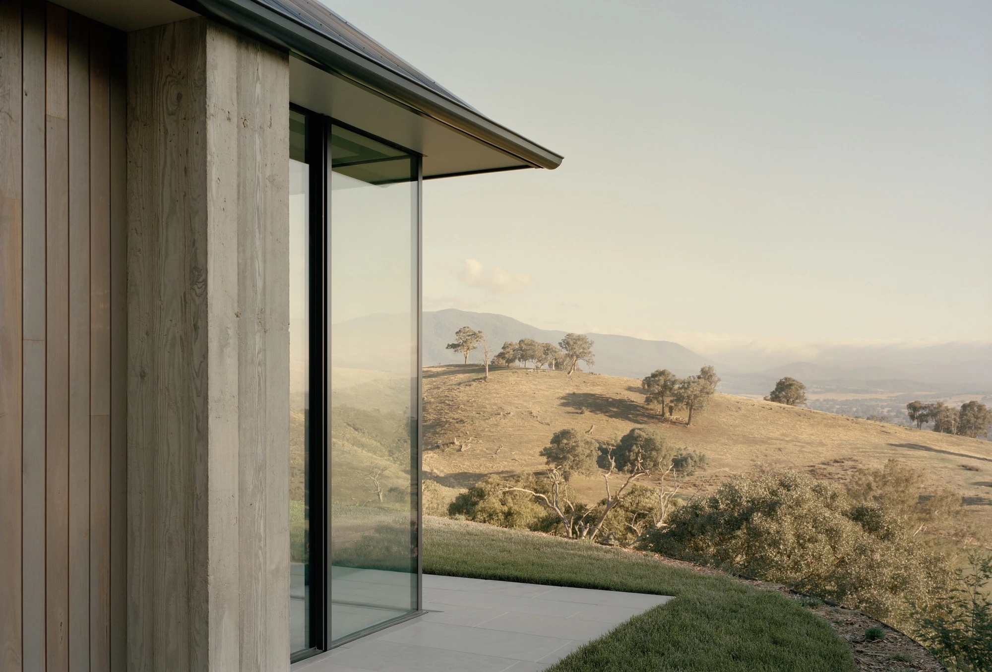 Cathedral House by JAK Architecture, photographed by Tasha Tylee — a sliding glass corner frames golden hills and scattered gum trees, timber cladding meeting a stone terrace at the building's edge.