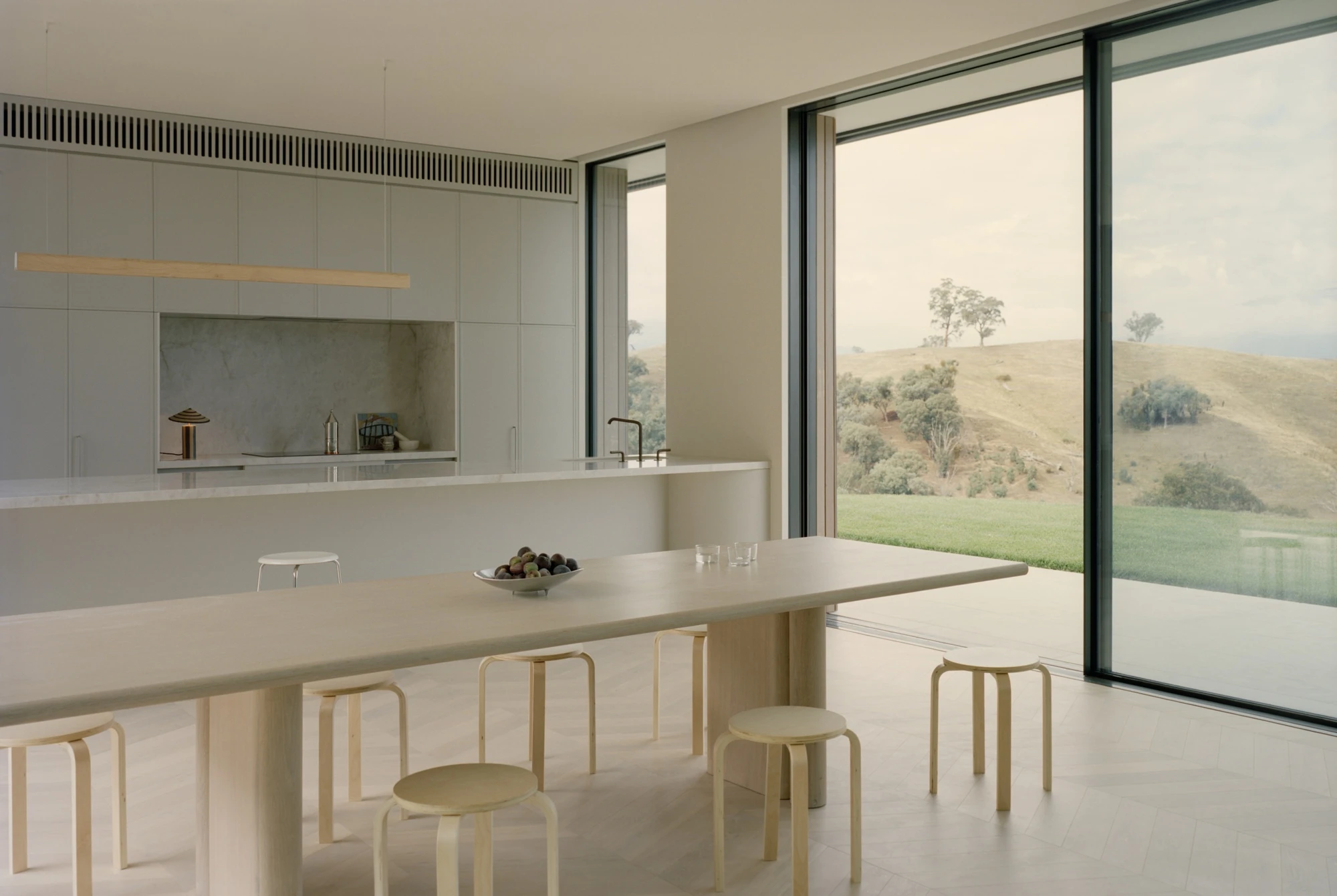 Cathedral House by JAK Architecture, photographed by Tasha Tylee — a pale open kitchen and dining room with a timber table, birch stools and a marble island, floor-to-ceiling glazing opening to the green paddock and hills.
