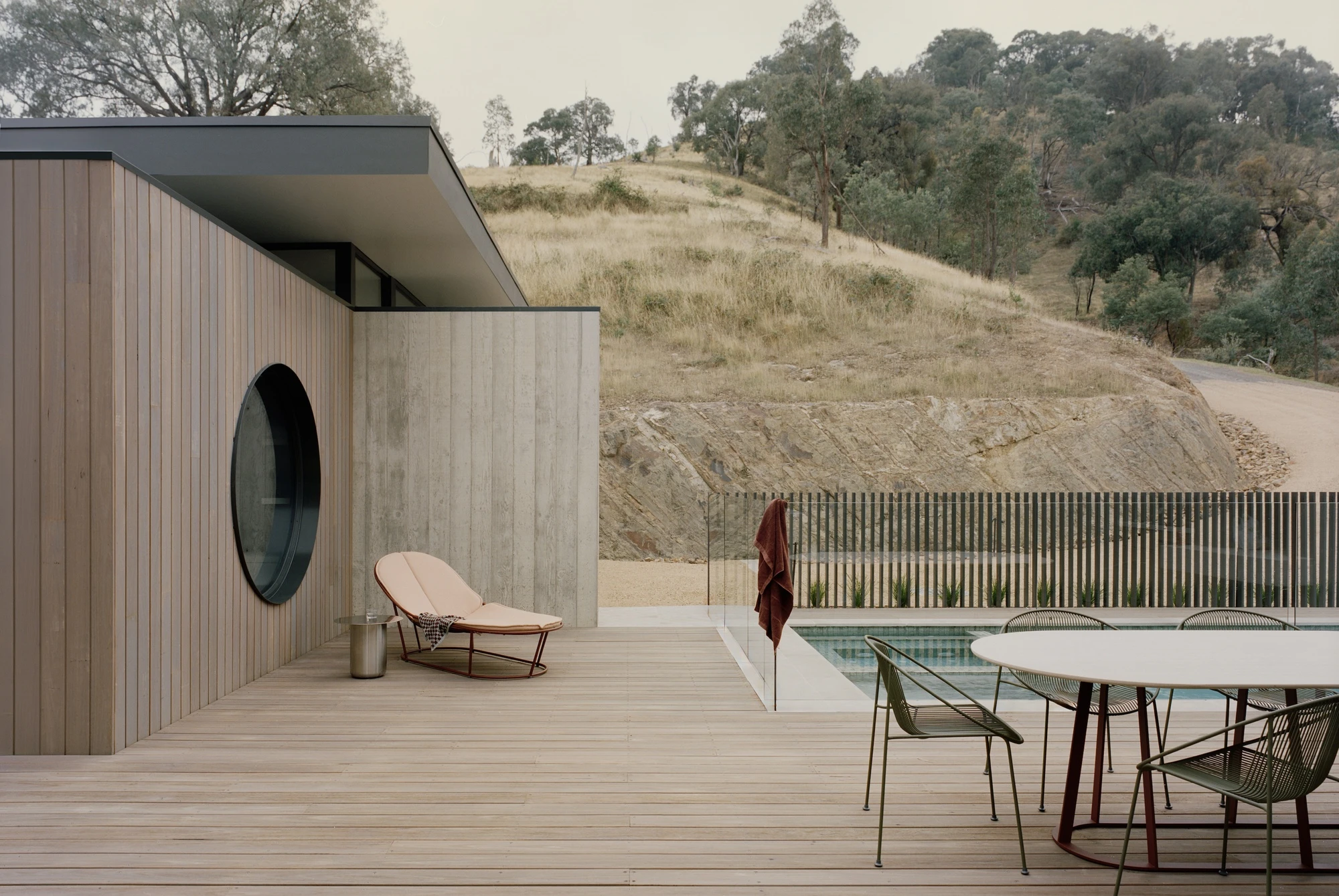 Cathedral House by JAK Architecture, photographed by Tasha Tylee — a pale timber deck with a leather chaise and dining setting, a black circular window in the concrete wall and the pool edged by a cut rock bank.