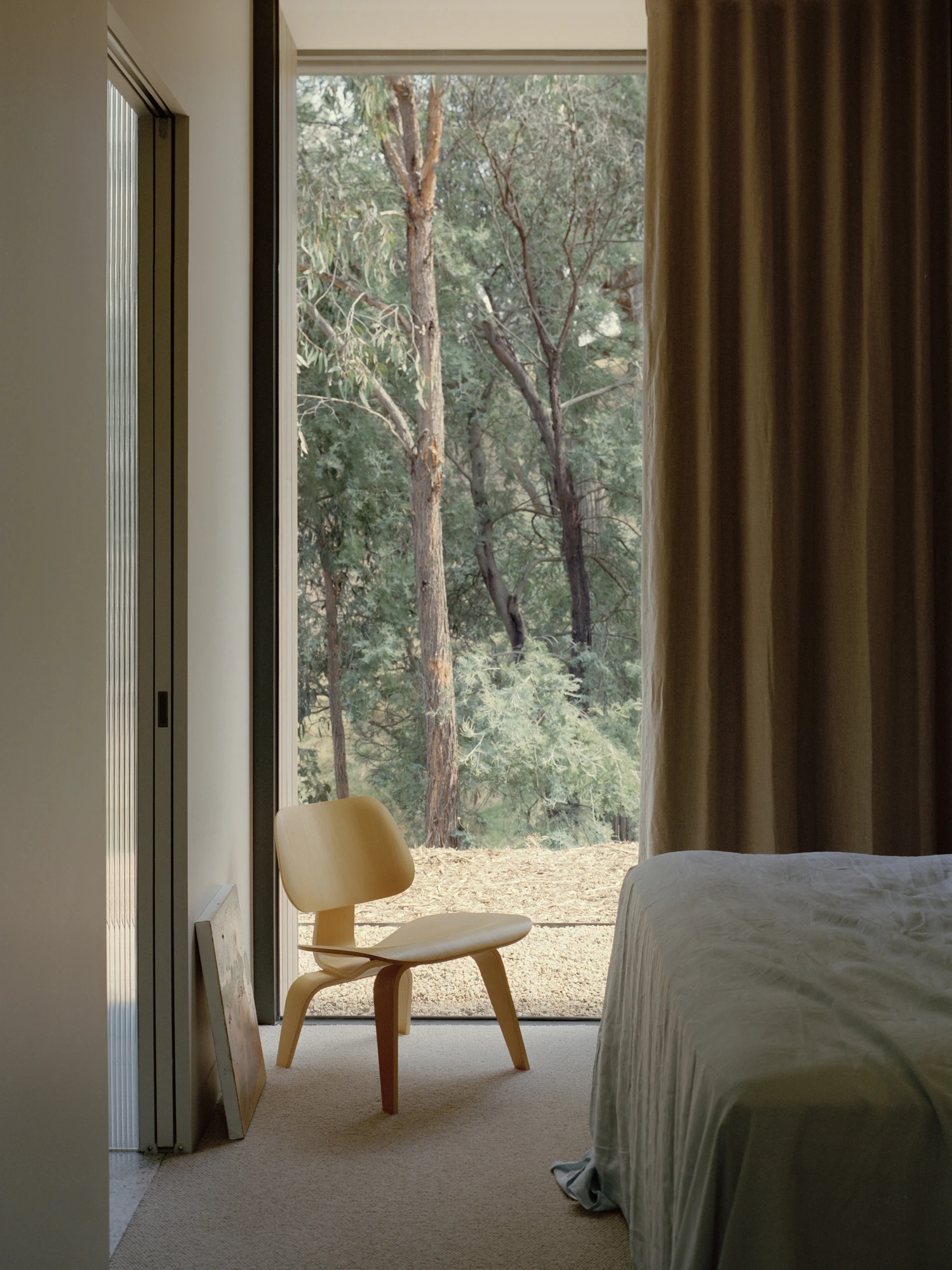Cathedral House by JAK Architecture, photographed by Tasha Tylee — a bedroom corner with a plywood lounge chair beside full-height glazing onto eucalypt bushland, a linen curtain drawn back and a grey-covered bed.