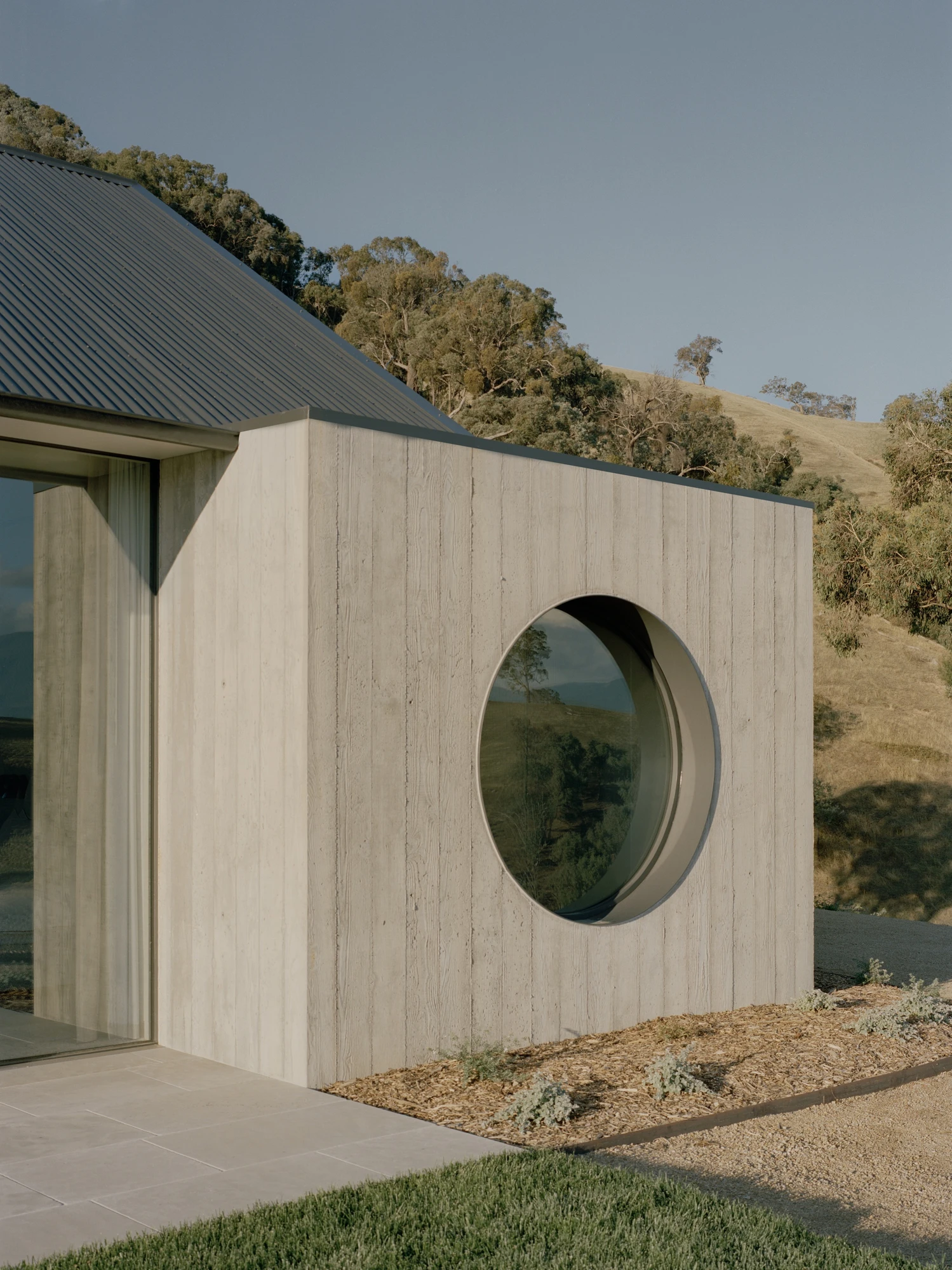 Cathedral House by JAK Architecture, photographed by Tasha Tylee — a board-formed concrete end wall holds a deep circular window that frames the ranges, the gabled roofline and dry hillside behind.