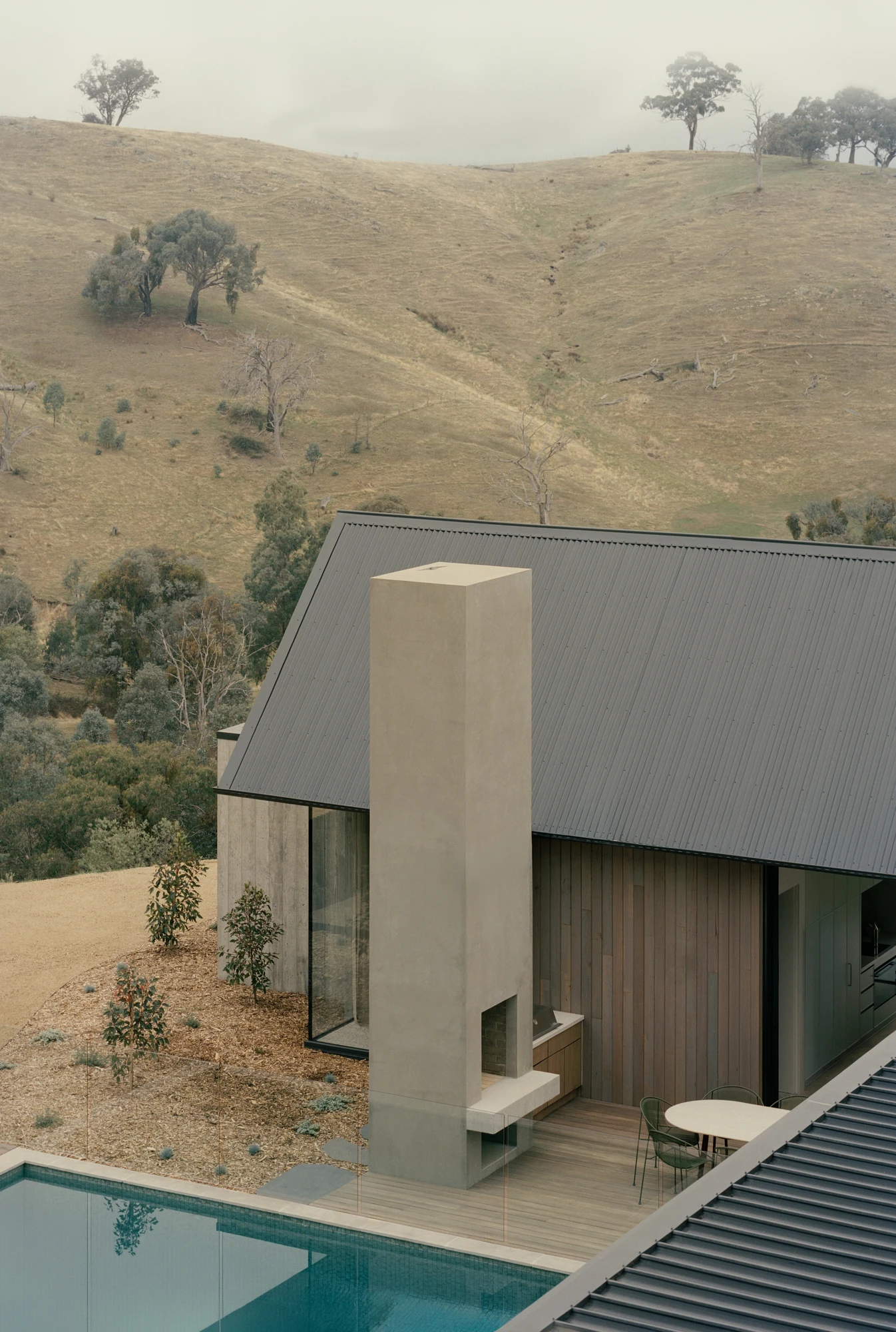 Cathedral House by JAK Architecture, photographed by Tasha Tylee — a tall plaster chimney rises beside timber cladding above the pool, the cut rock face and dry hills climbing behind.