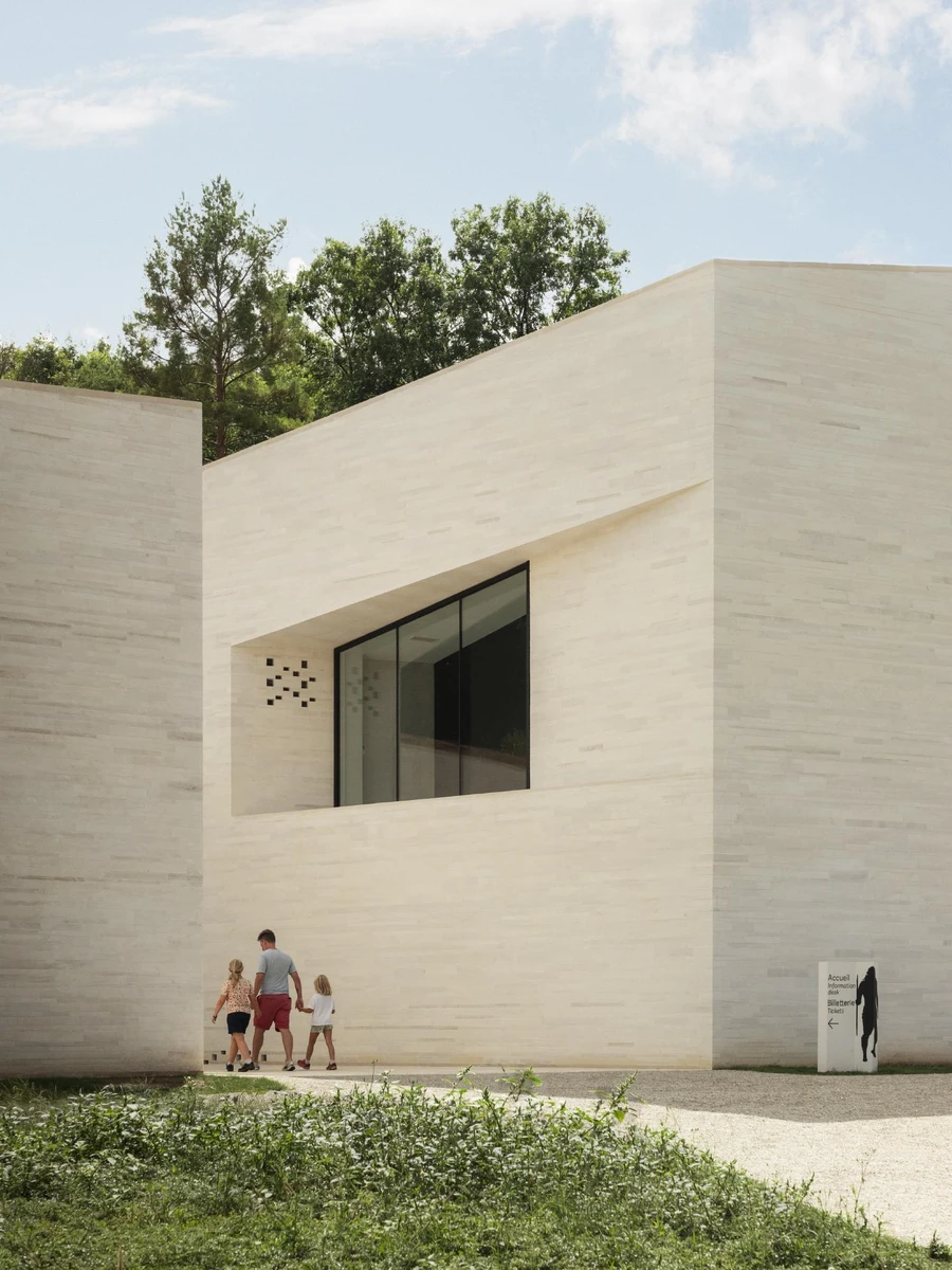 Neanderthal Museum, La Chapelle-aux-Saints by W-Architectures, photographed by Cyrille Weiner — a family walks a gravel path toward a layered-concrete corner with a long horizontal window cut into it.