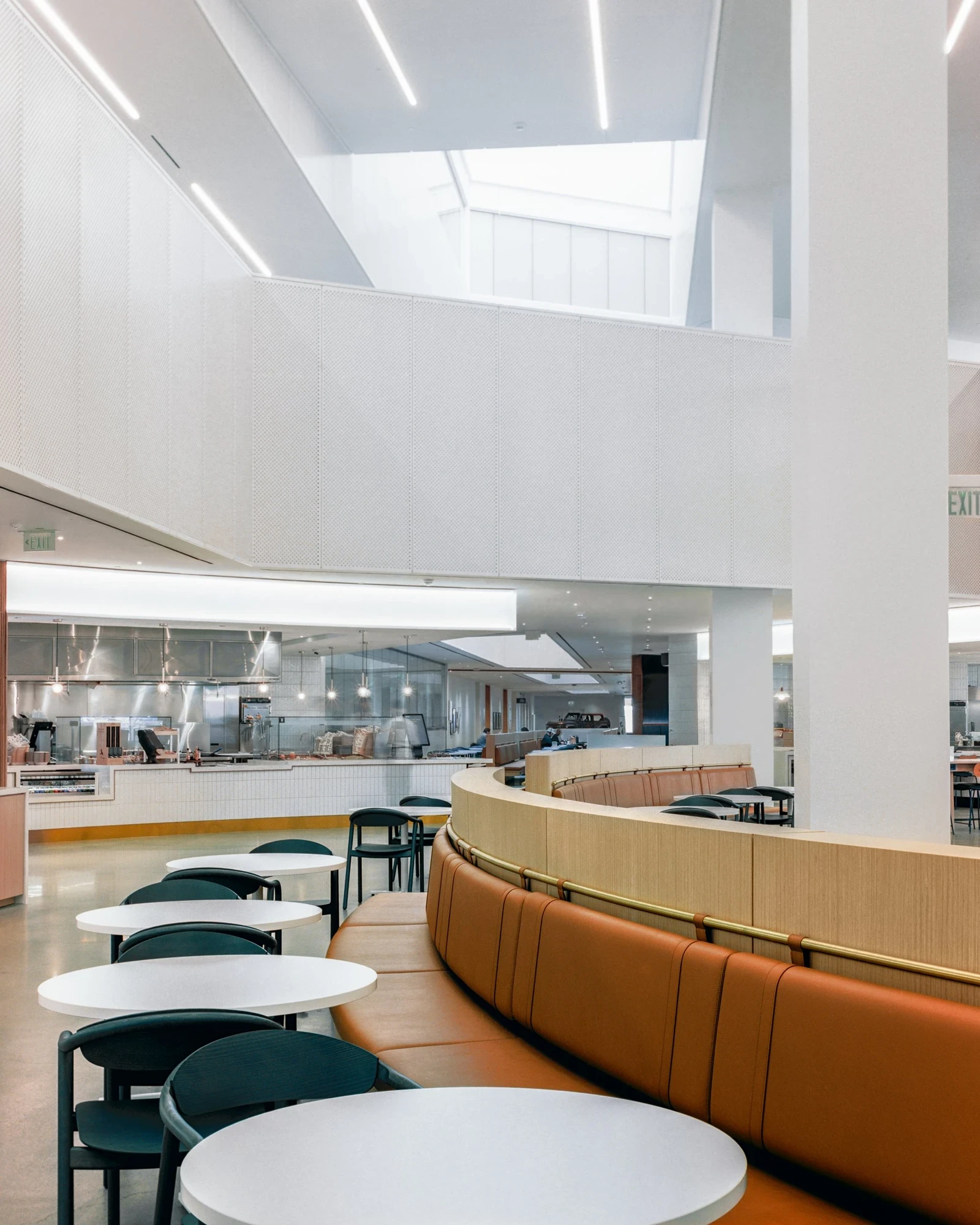 Ford World Headquarters by Snøhetta, photographed by Aaron Feliciano — a curved tan-leather and oak banquette wraps the food hall seating beneath the white perforated shell, the open kitchen beyond.
