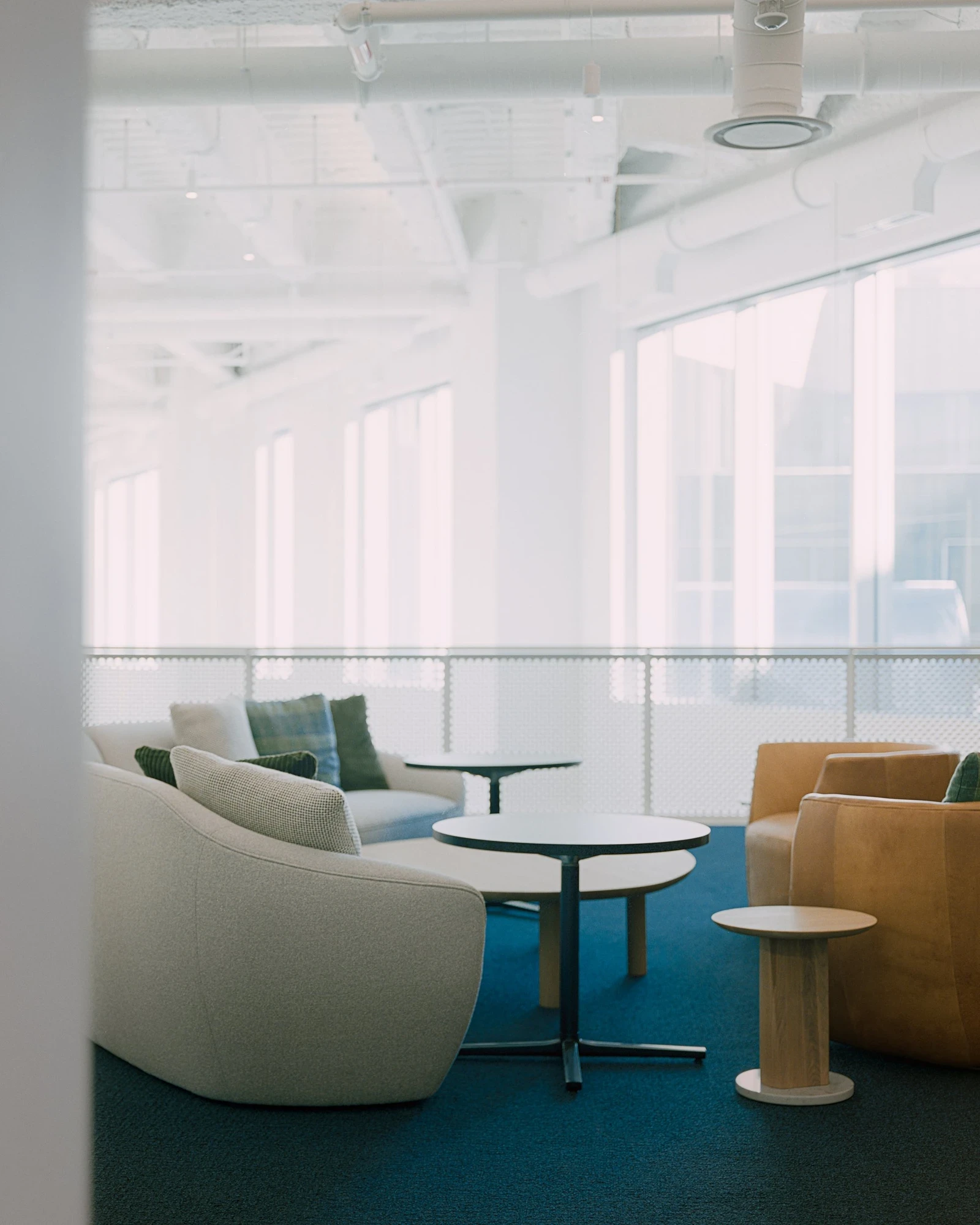 Ford World Headquarters by Snøhetta, photographed by Aaron Feliciano — an upper-floor lounge with curved sofas on teal carpet, ducts and structure left exposed and painted white overhead.