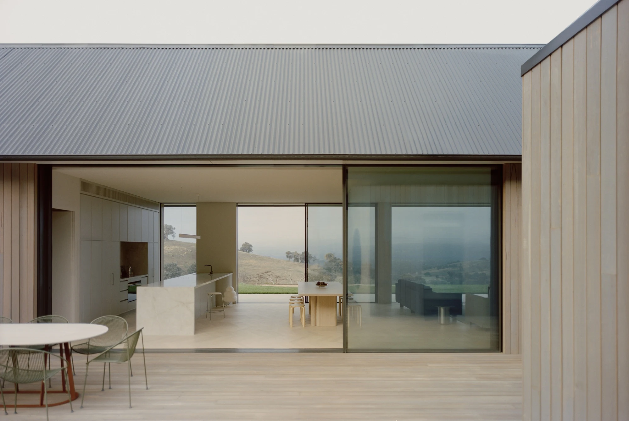 Cathedral House by JAK Architecture, photographed by Tasha Tylee — a green velvet corner sofa faces a frameless glass wall that opens onto rolling hills and a distant valley.