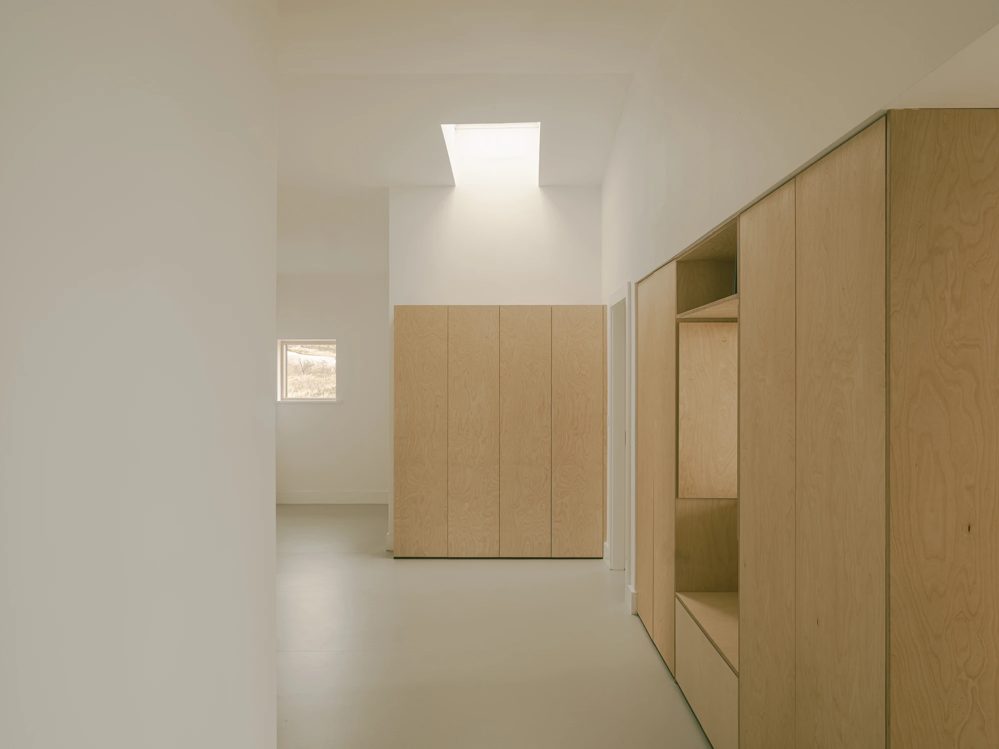 Cruit Island by Pasparakis Friel, photographed by Peter Molloy — a top-lit hallway lined with birch plywood storage, a small window framing wind-swept grass at its end.