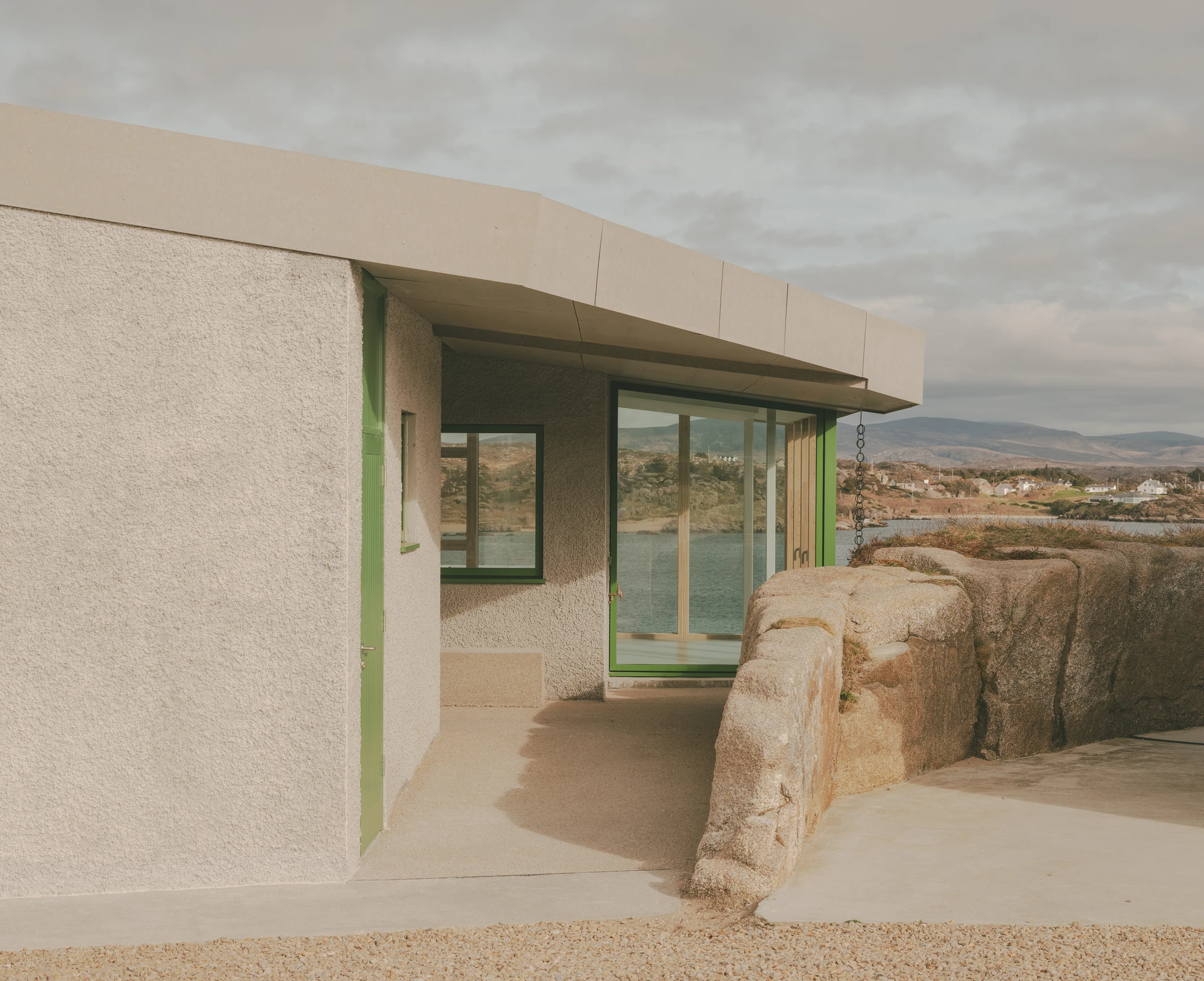 Cruit Island by Pasparakis Friel, photographed by Peter Molloy — a fibre cement roof curves over a rendered corner where green-framed glazing meets a boulder abutting the wall.