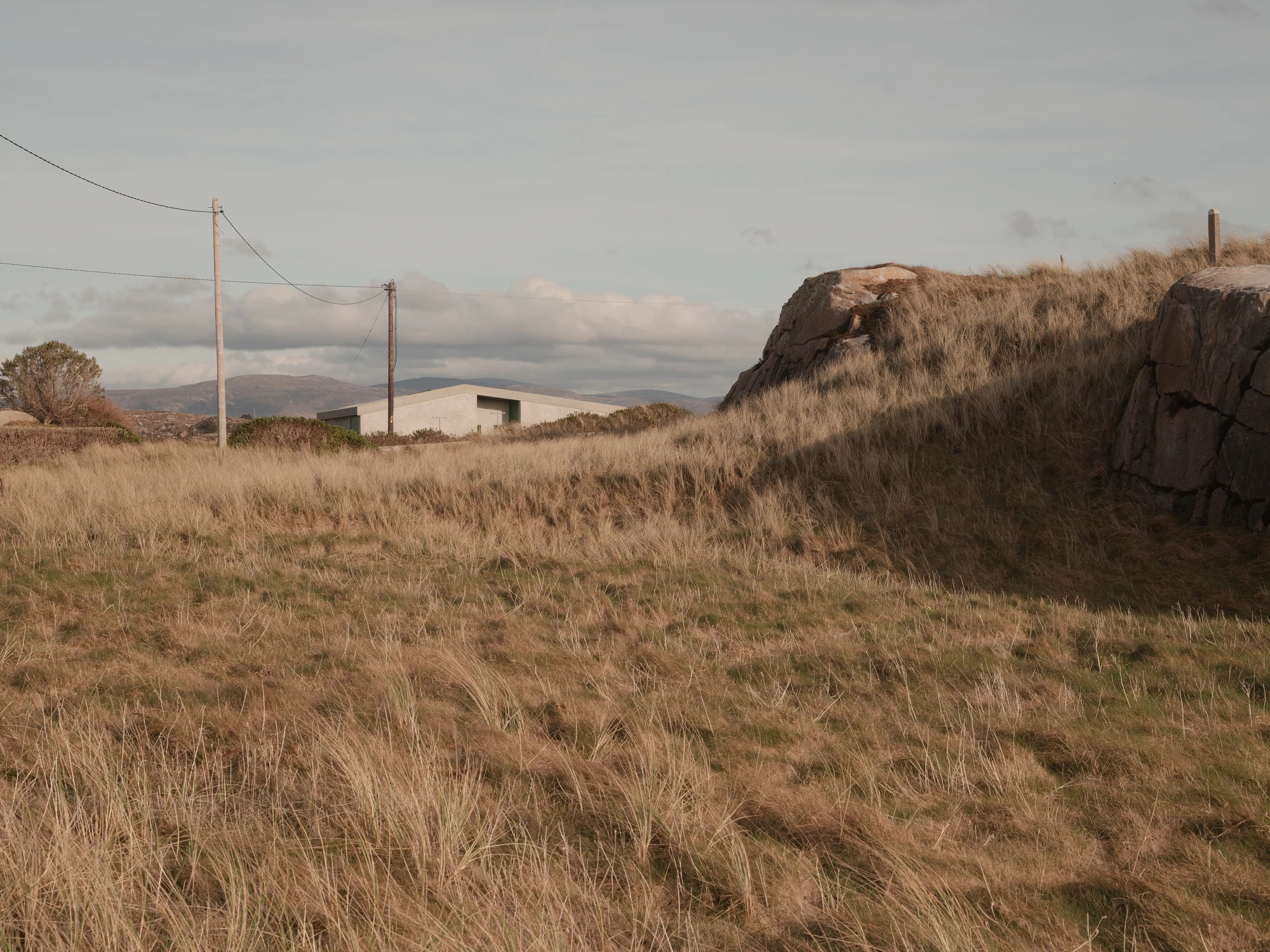 Cruit Island by Pasparakis Friel, photographed by Peter Molloy — the pale render mass of the house sits low behind wind-swept grass and granite outcrops, Errigal Mountain faint on the horizon.