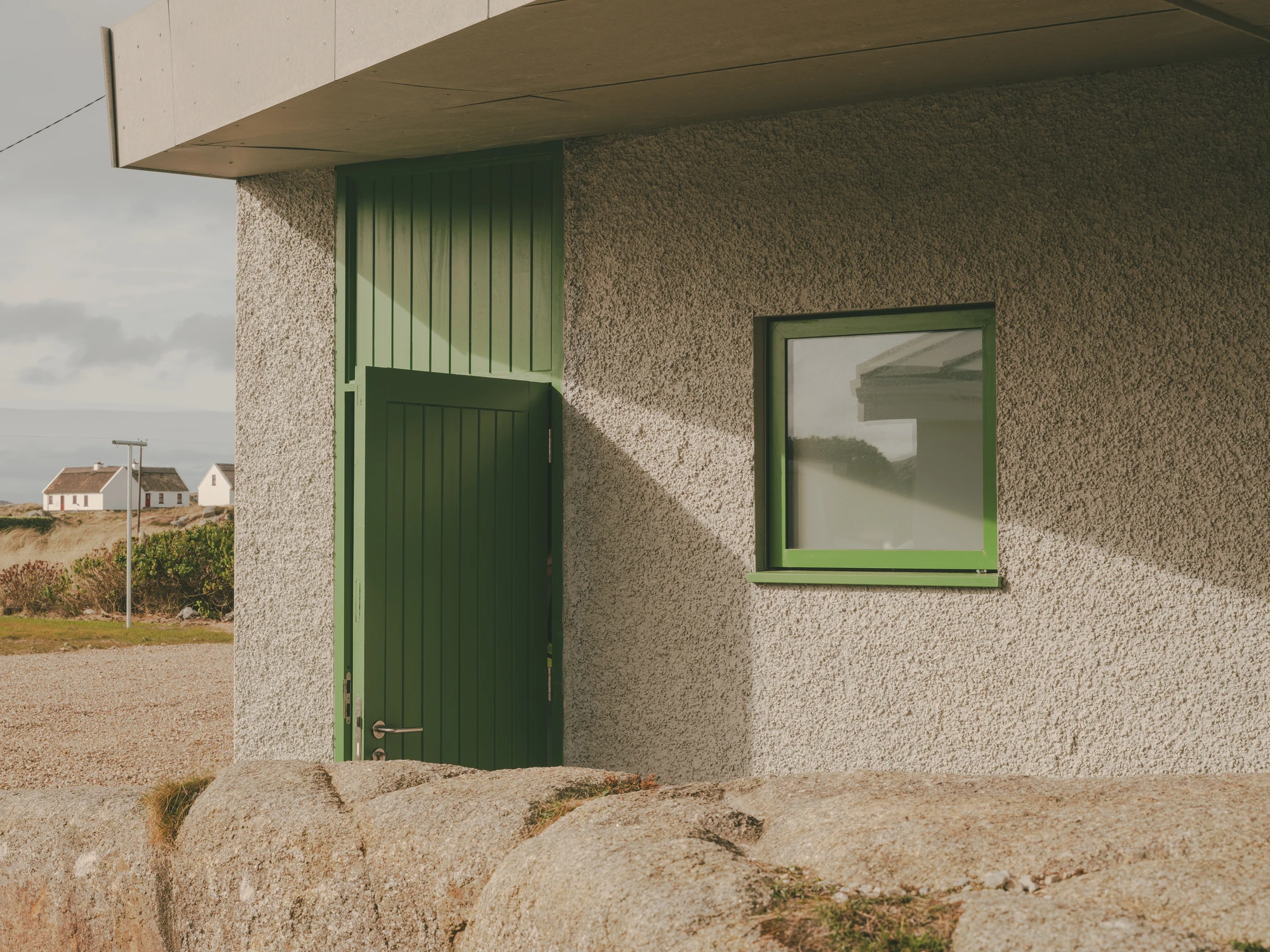 Cruit Island by Pasparakis Friel, photographed by Peter Molloy — a green timber door and a green-framed square window set into pebbled roughcast render, granite rock rising in the foreground.