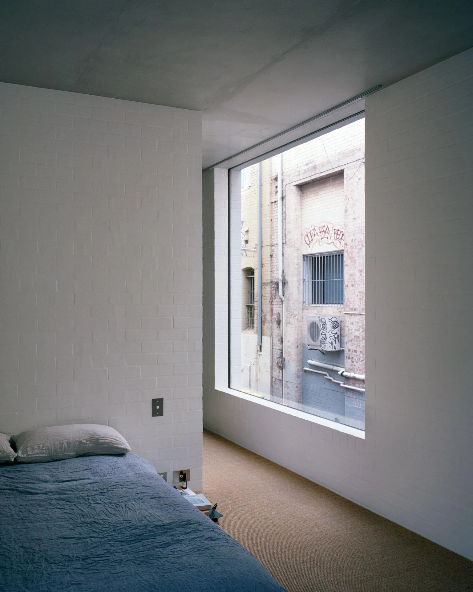 House next to Taylor Square by Anthony Gill Architects, photographed by Rory Gardiner with Colby Vexler — Bedroom in white painted brick, a corner window cut to frame the graffiti-tagged facade across the lane.