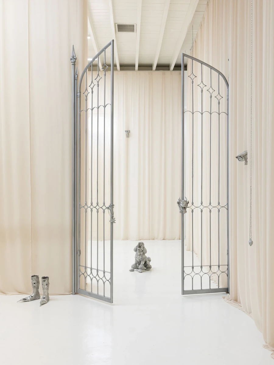You Can't Stop the World from Being Bad by Andrea Ferrero — Lima, Peru: the ornate arched double gate open wide, framing a seated silver poodle beyond, cast knight's boots at left and a heart-lensed camera on the curtain.