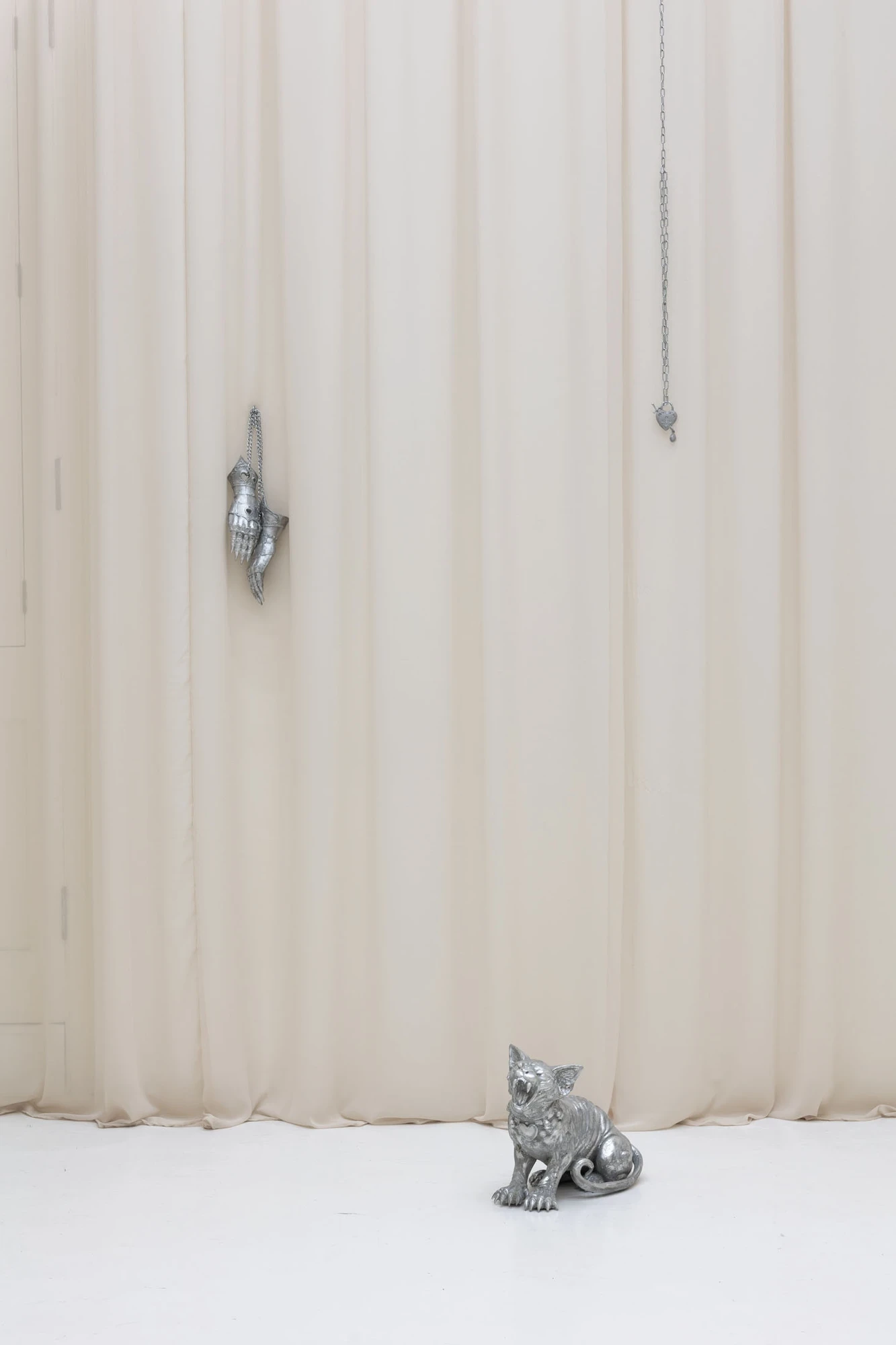 You Can't Stop the World from Being Bad by Andrea Ferrero, photographed by Hector Delgado — an armoured gauntlet and a love padlock hanging on chains from the curtain, a small silver cat sculpture seated on the floor below.