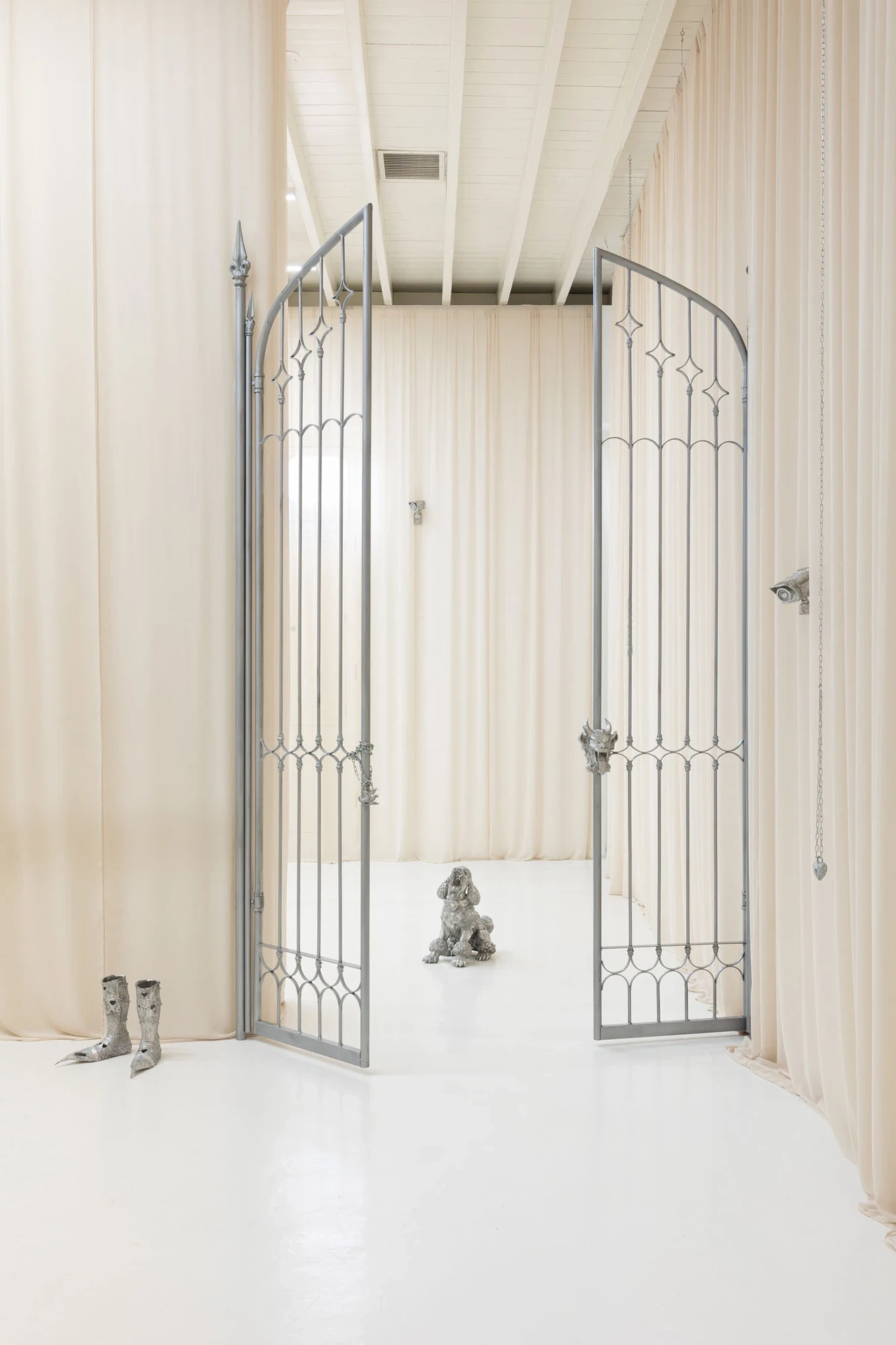 You Can't Stop the World from Being Bad by Andrea Ferrero, photographed by Hector Delgado — the ornate arched double gate open wide, framing a seated silver poodle beyond, cast knight's boots at left and a heart-lensed camera at right.