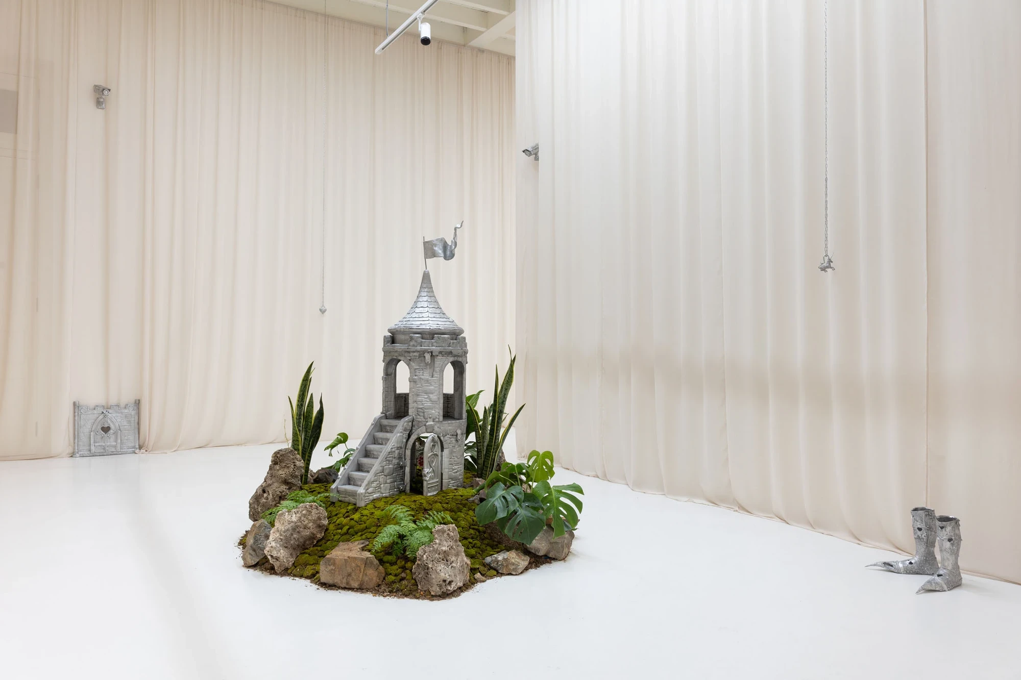 You Can't Stop the World from Being Bad by Andrea Ferrero, photographed by Hector Delgado — the moss castle island with its tower and rocks at the centre of the room, cast silver boots to the right and a heart-cut wall fragment far left.