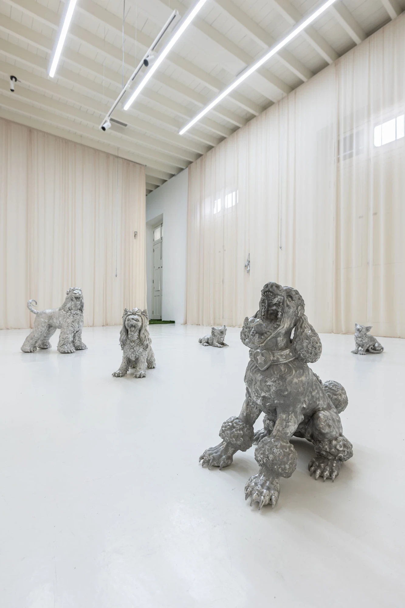 You Can't Stop the World from Being Bad by Andrea Ferrero, photographed by Hector Delgado — silver cast dog sculptures of assorted breeds and sizes distributed across a white gallery floor, sheer curtains veiling the walls behind them.