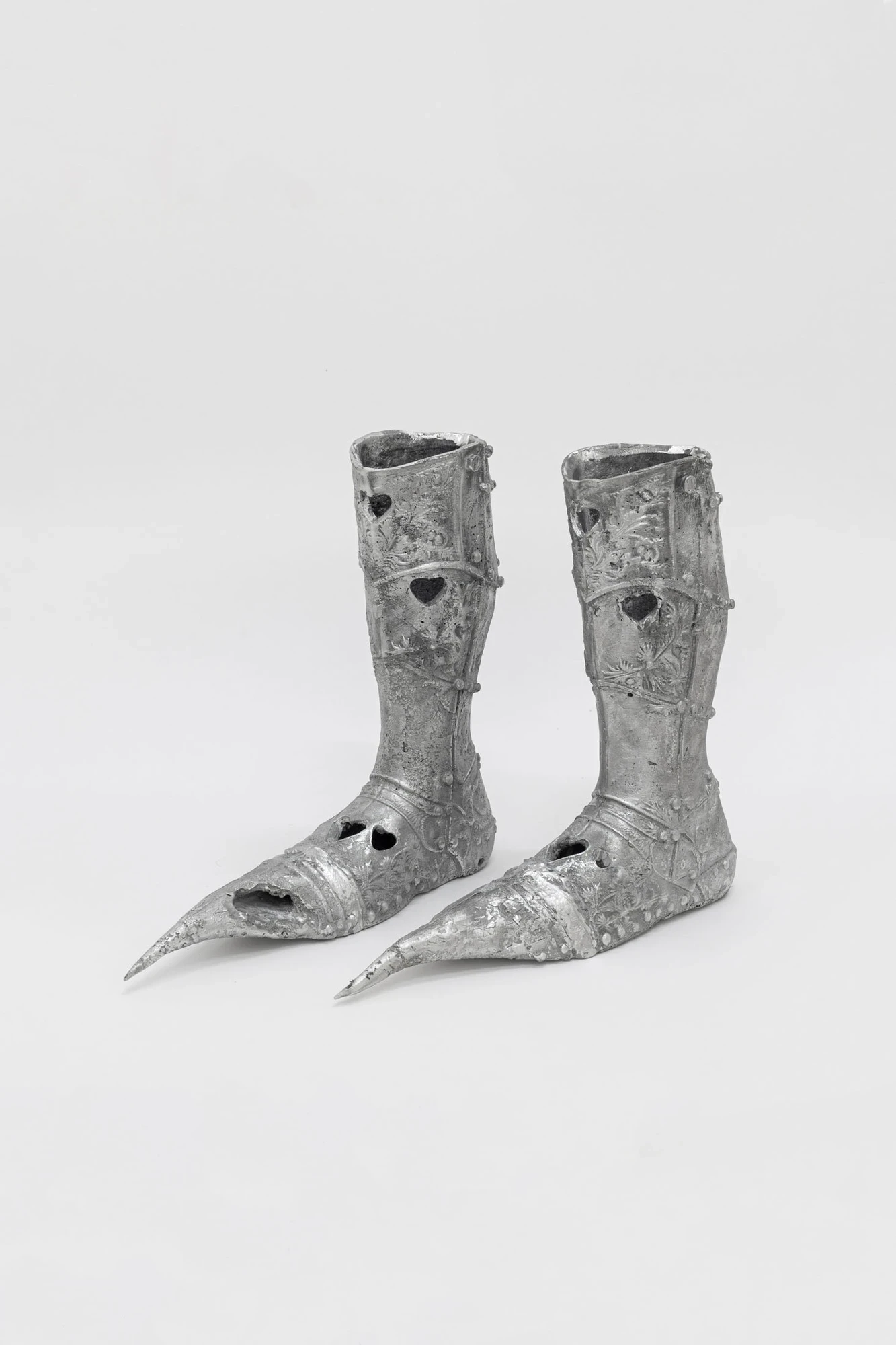 You Can't Stop the World from Being Bad by Andrea Ferrero, photographed by Hector Delgado — a black-and-white studio shot of a pair of cast silver sabatons, pointed mediaeval armour boots perforated with hearts and engraved with flowers.