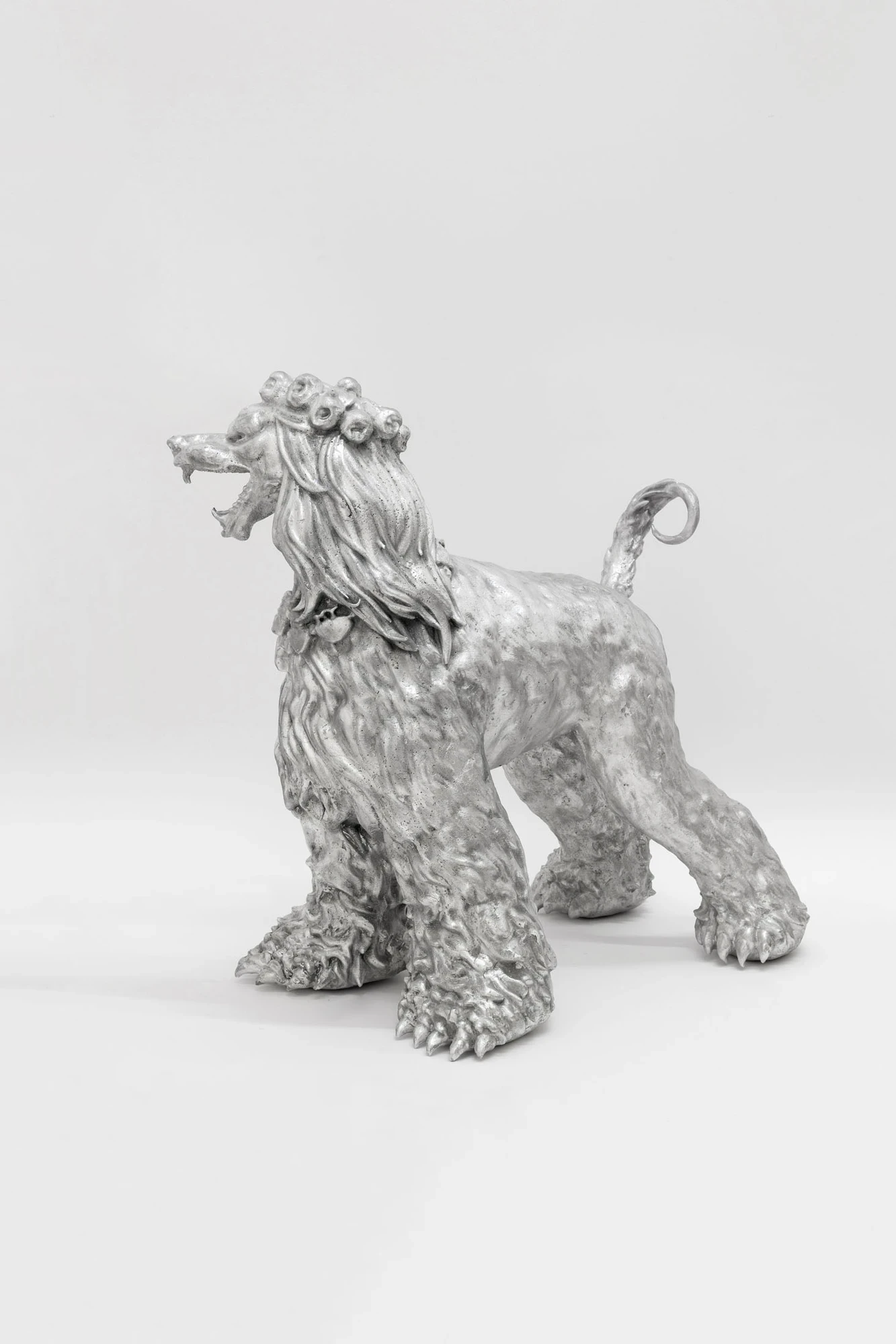 You Can't Stop the World from Being Bad by Andrea Ferrero, photographed by Hector Delgado — a black-and-white studio shot of a silver Afghan hound sculpture with a crown, bell collar and open mouth, seen from a rear quarter angle.