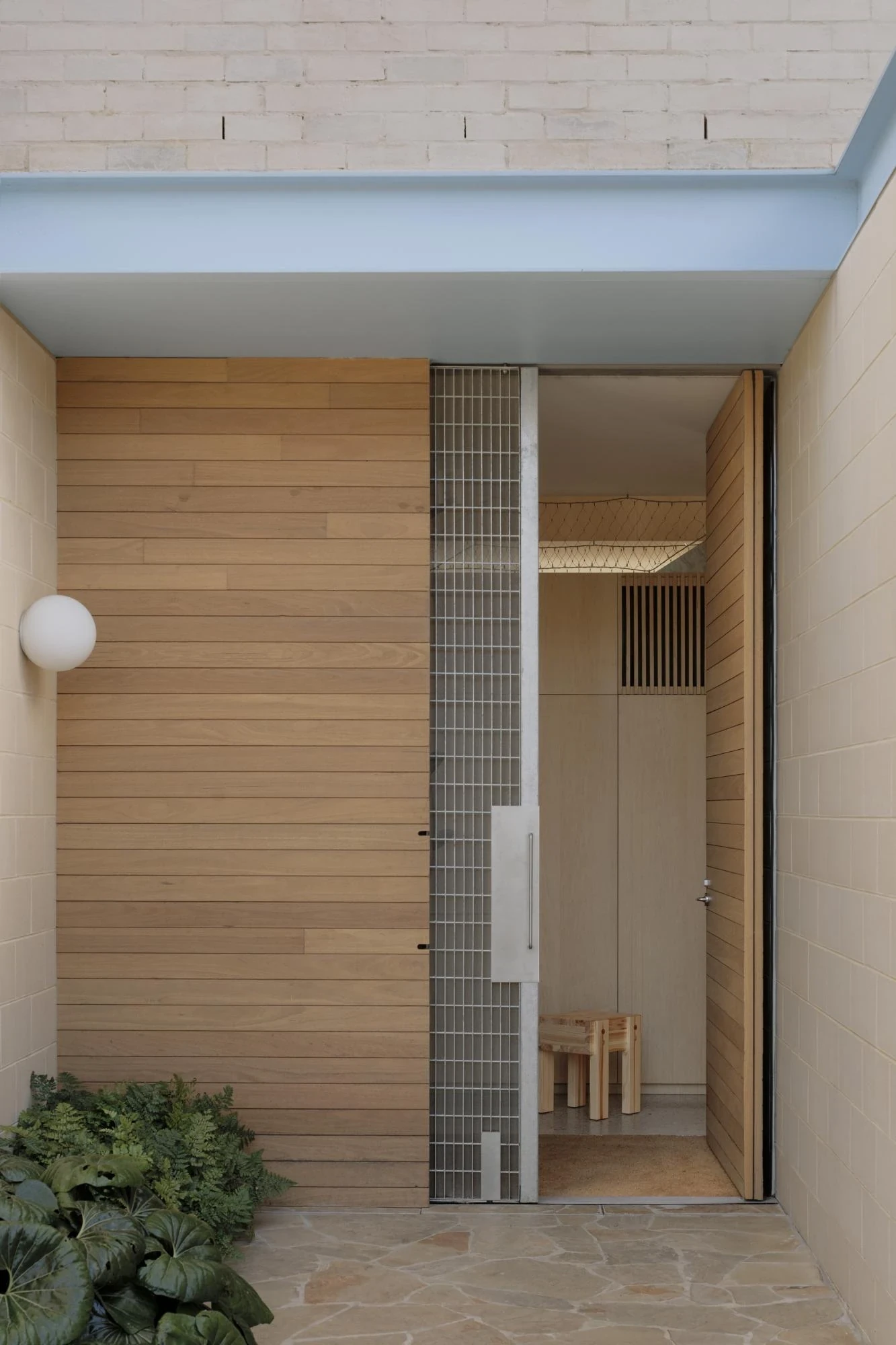 A Courtyard House by Office MIJI, photographed by Ben Hosking — an entry of horizontal timber cladding and a stainless grille door beside a globe wall light, stone paving underfoot.