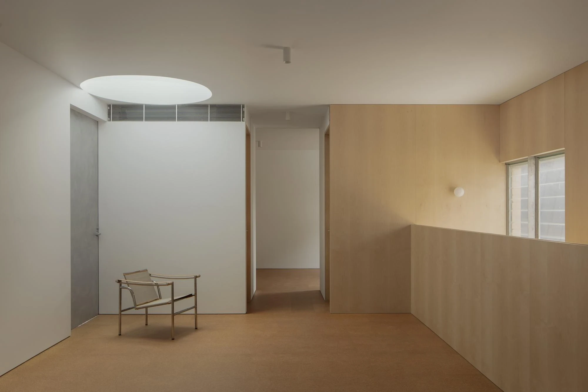 A Courtyard House by Office MIJI, photographed by Ben Hosking — an upper landing with a circular oculus and cork floor, a wire-and-leather chair against a timber-veneer wall.