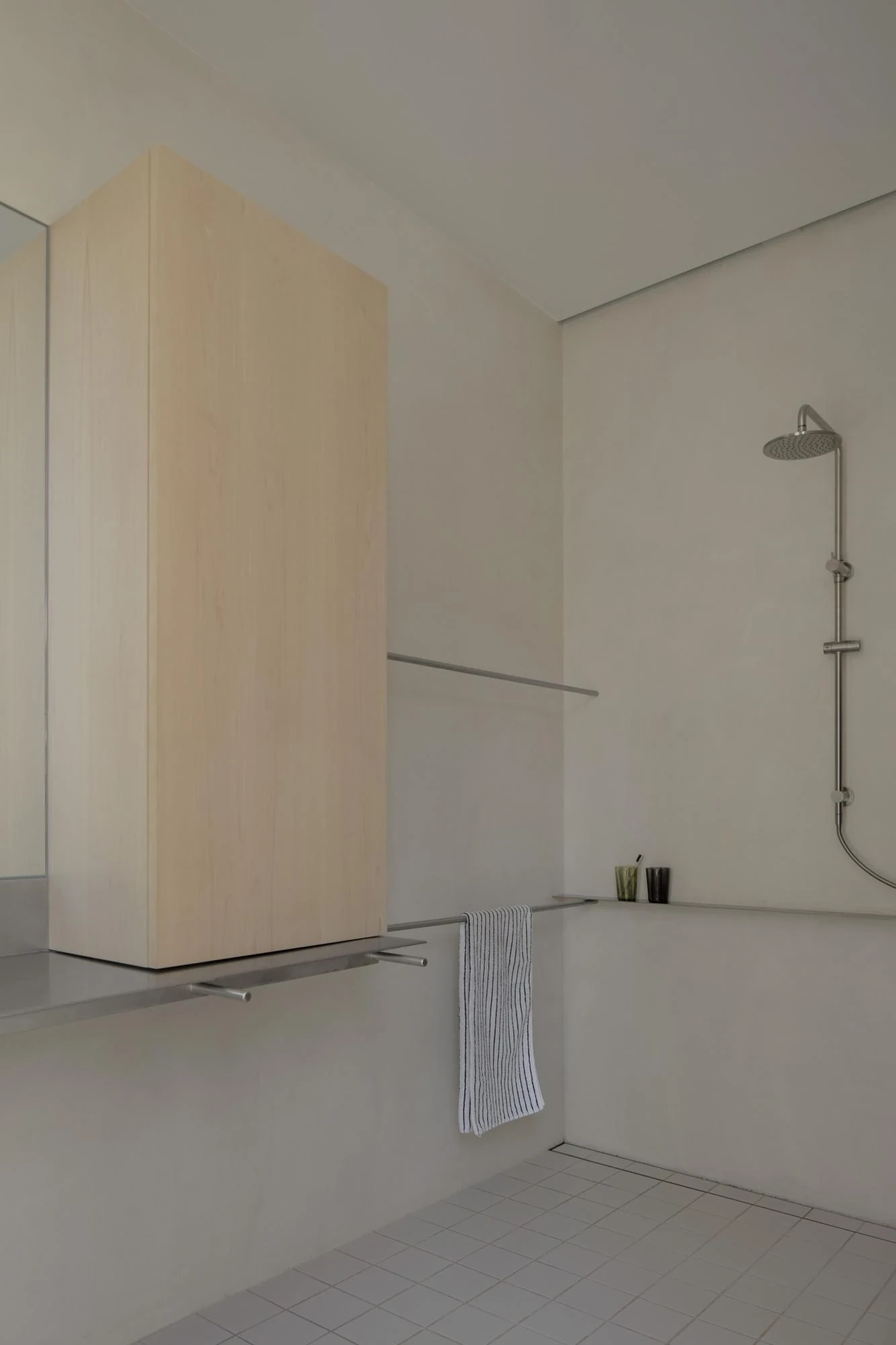 A Courtyard House by Office MIJI, photographed by Ben Hosking — a rendered bathroom with a stainless rail vanity, a wall shower and pale ash cabinet over white floor tile.