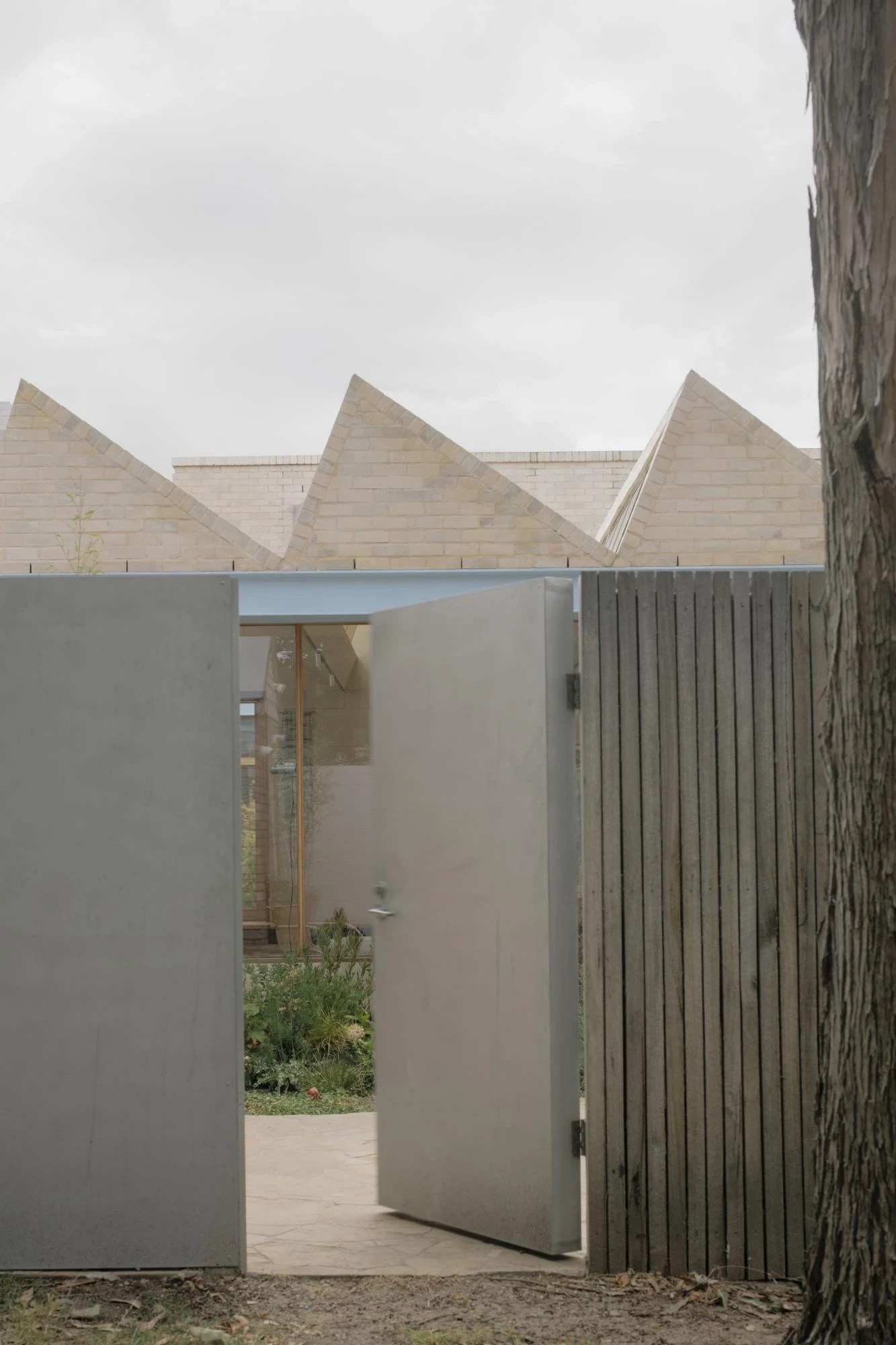 A Courtyard House by Office MIJI, photographed by Ben Hosking — a pivoting concrete gate opens between fence and wall toward a garden path, the sawtooth brick roofline overhead.