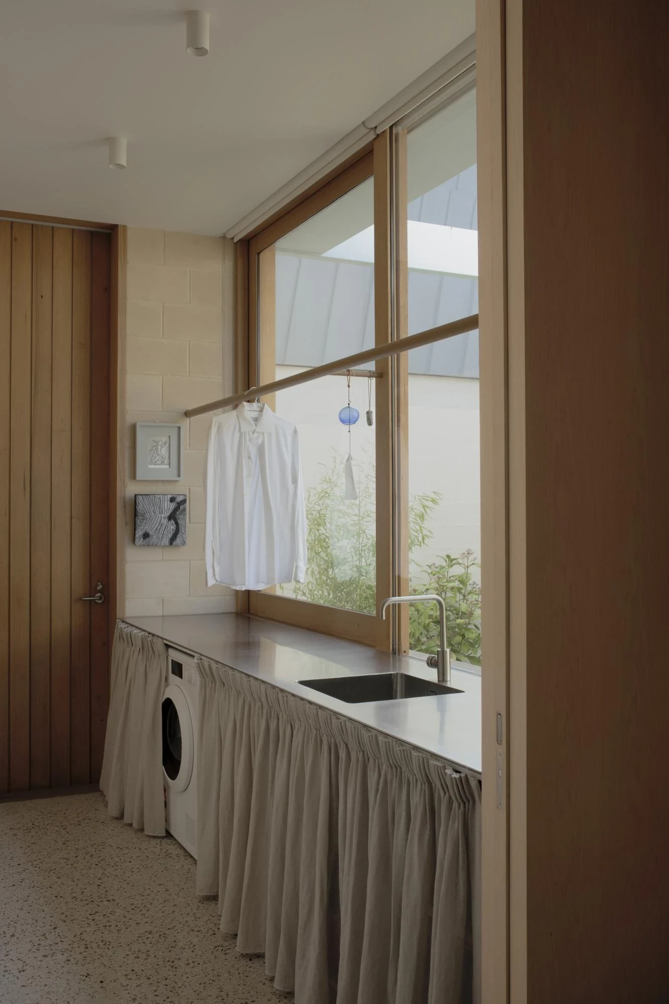 A Courtyard House by Office MIJI, photographed by Ben Hosking — the laundry with a machine hidden behind a linen skirt below a window, a white shirt hung to dry on a timber rail.