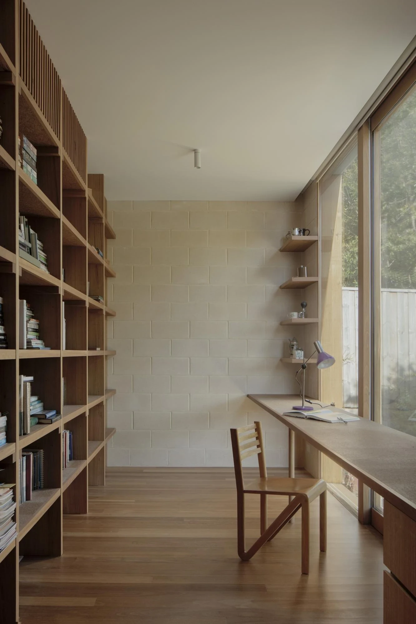 A Courtyard House by Office MIJI, photographed by Ben Hosking — the study wall of timber book stacks facing a long desk pushed to full-height glazing, a purple lamp on the desk.