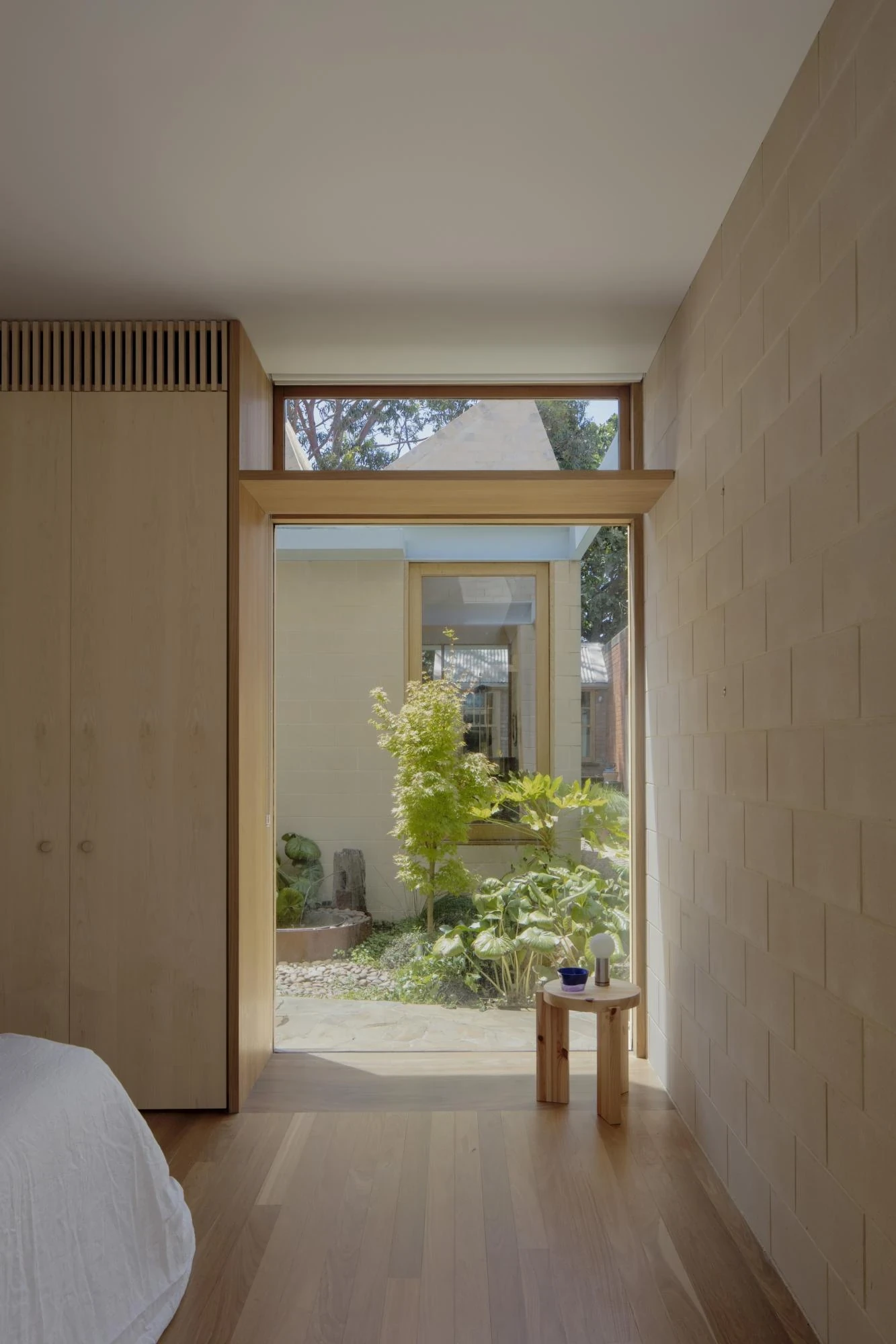 A Courtyard House by Office MIJI, photographed by Ben Hosking — a bedroom of ash joinery opening onto a small stone garden with a maple, a pine stool at the threshold.