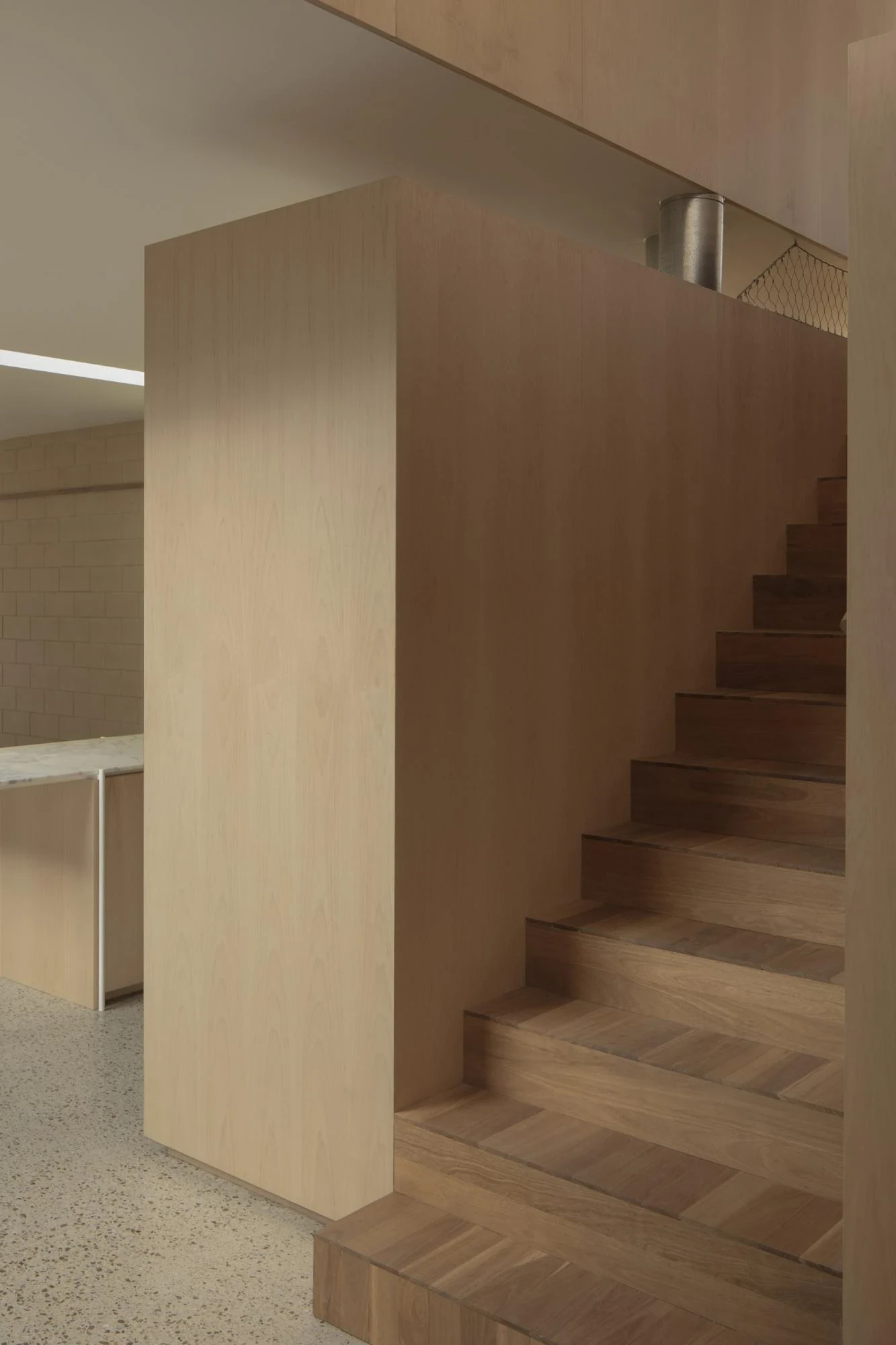 A Courtyard House by Office MIJI, photographed by Ben Hosking — a spotted-gum stair rising beside a smooth ash-veneer wall toward the kitchen on terrazzo.