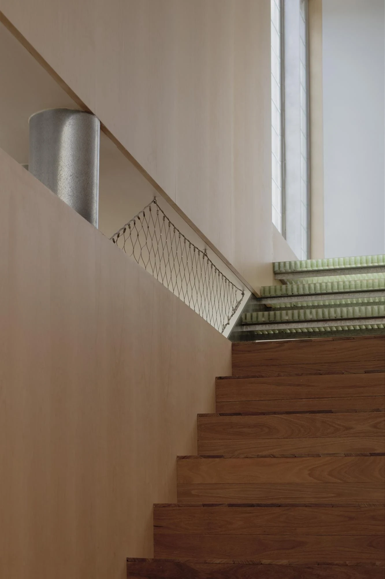 A Courtyard House by Office MIJI, photographed by Ben Hosking — the stair turning below a green glass-block skylight, a galvanised downpipe and wire-mesh balustrade catching the light.