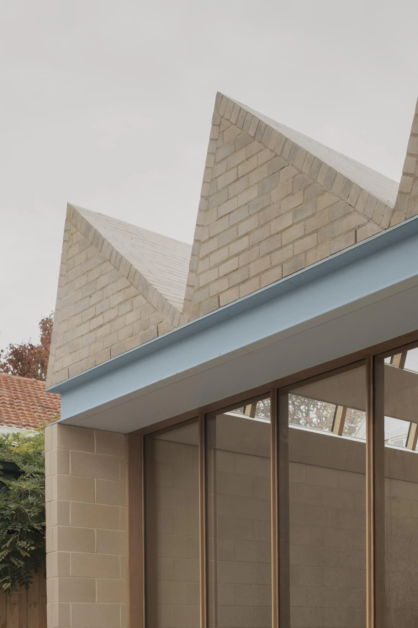 A Courtyard House by Office MIJI, photographed by Ben Hosking — a close view of two cream-brick gables meeting a pale blue steel fascia above a cement-block wall and full-height glazing.