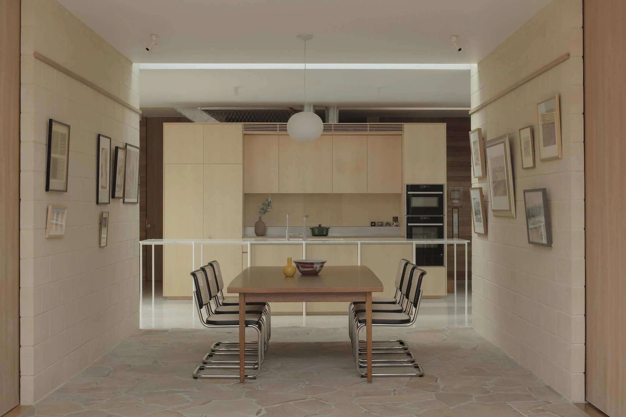 A Courtyard House by Office MIJI, photographed by Ben Hosking — the dining table framed between cement-block walls hung with prints, the ash kitchen beyond over crazy-paving stone.
