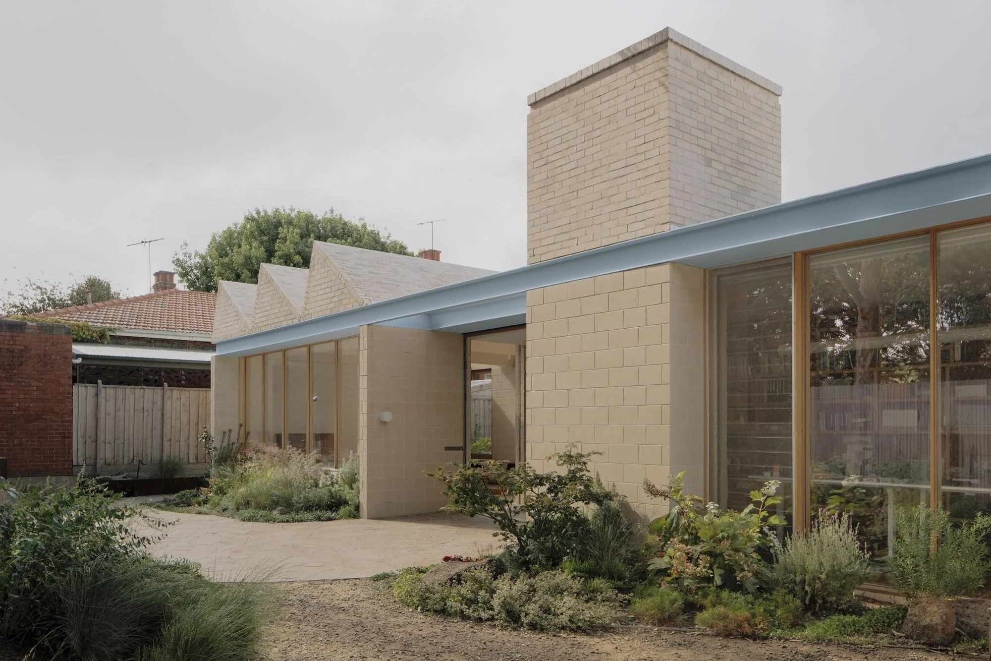 A Courtyard House by Office MIJI, photographed by Ben Hosking — the garden courtyard with planted beds and crazy-paving stone, pale blue steel band and brick chimney reflecting in the glass.
