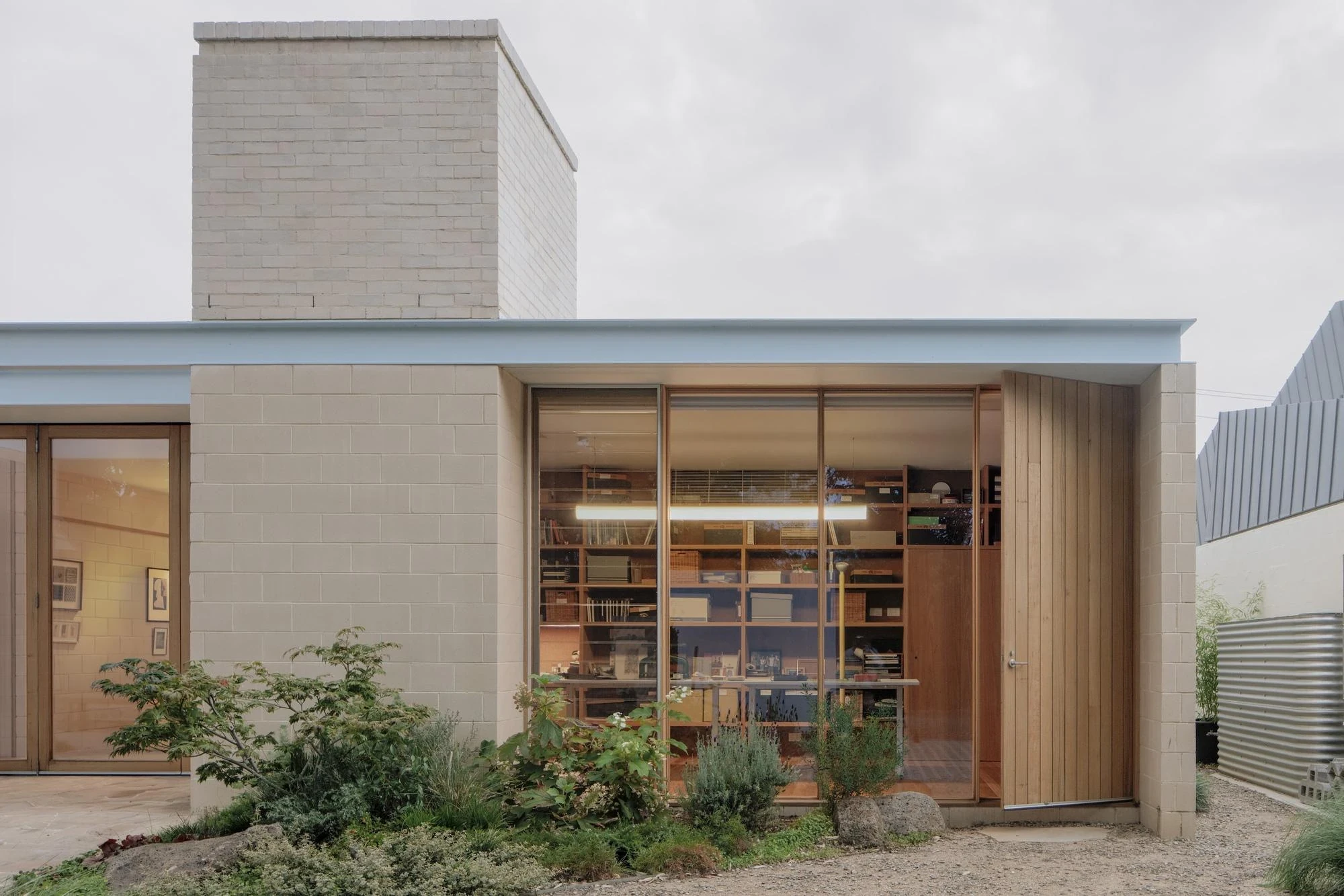 A Courtyard House by Office MIJI, photographed by Ben Hosking — the study seen from the garden through full-height glazing, book stacks glowing under a linear light below the brick chimney.