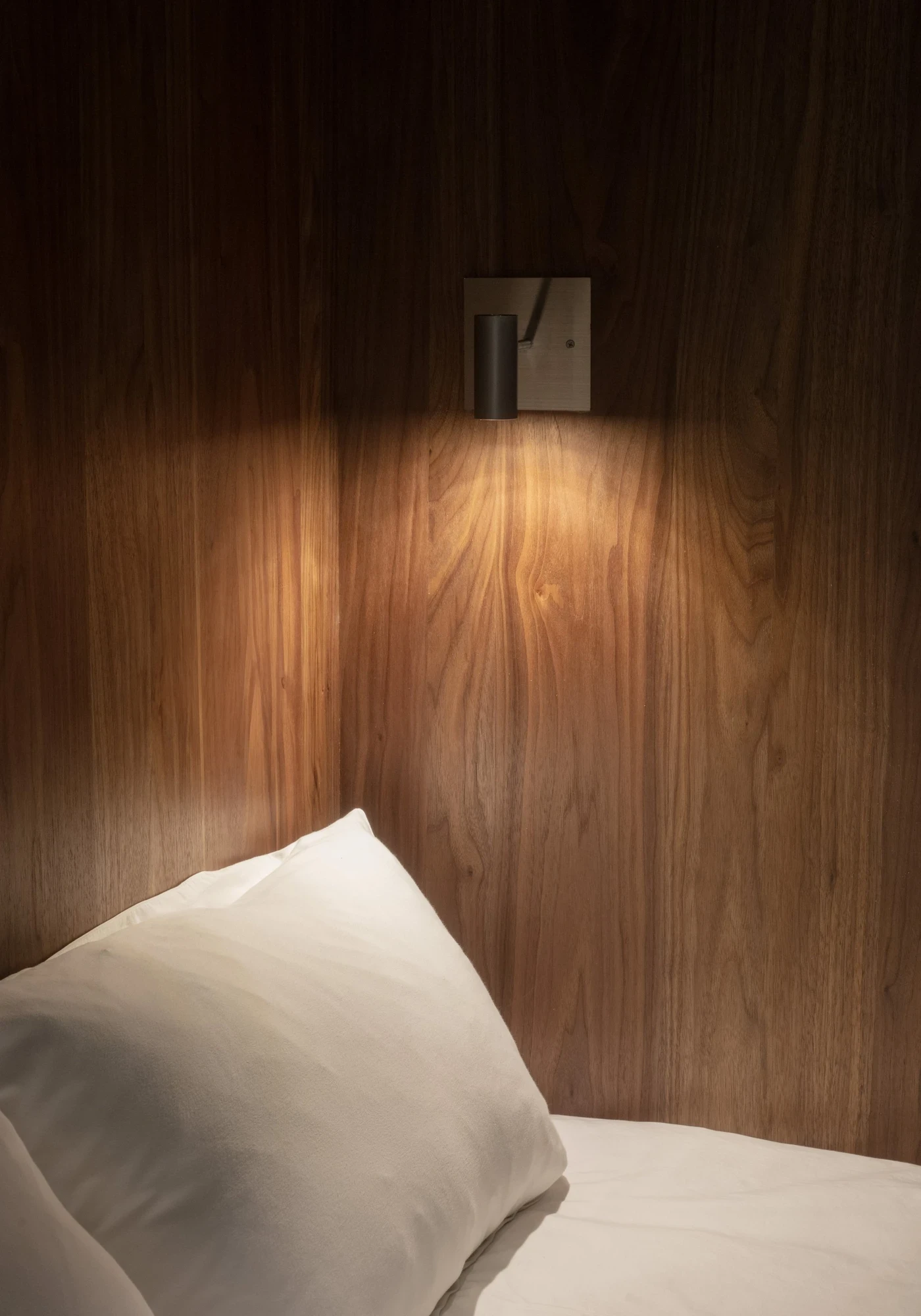 Ximen Apartment by unprofessional.studio, photographed by unprofessional.studio: a brushed chrome sconce pools warm amber light across the walnut wall grain above a white pillow.