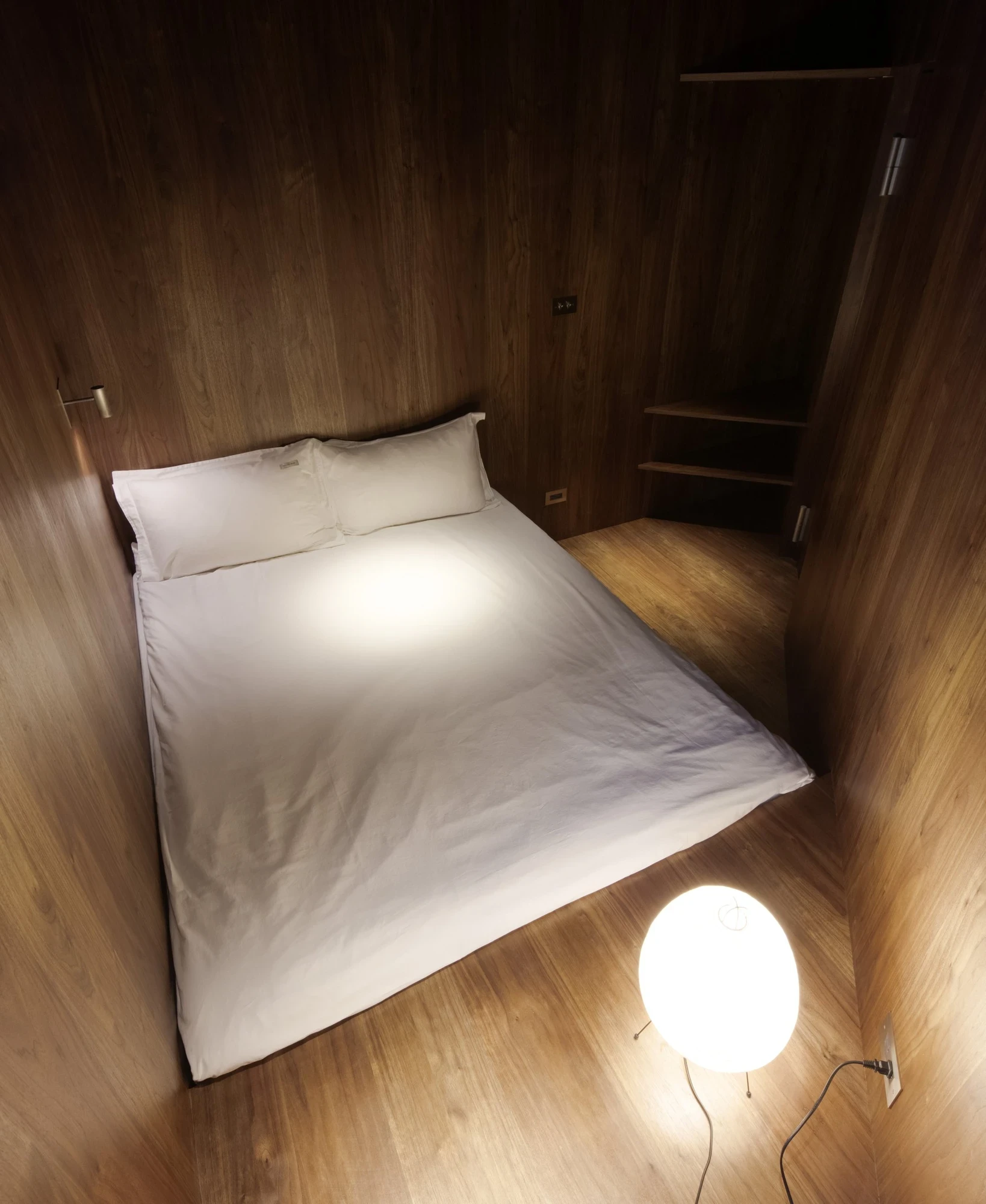 Ximen Apartment by unprofessional.studio, photographed by unprofessional.studio: the walnut-lined bedroom closes around a low white bed like a hull, a round globe lamp glowing on the oak floor.