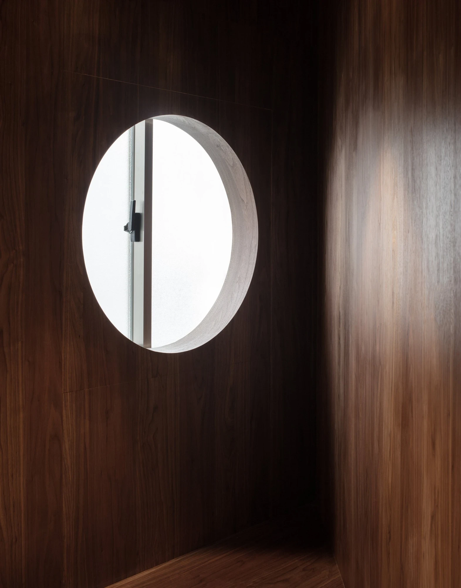 Ximen Apartment by unprofessional.studio, photographed by unprofessional.studio: a circular porthole window set into the dark walnut wall pours daylight across the opposite timber surface.