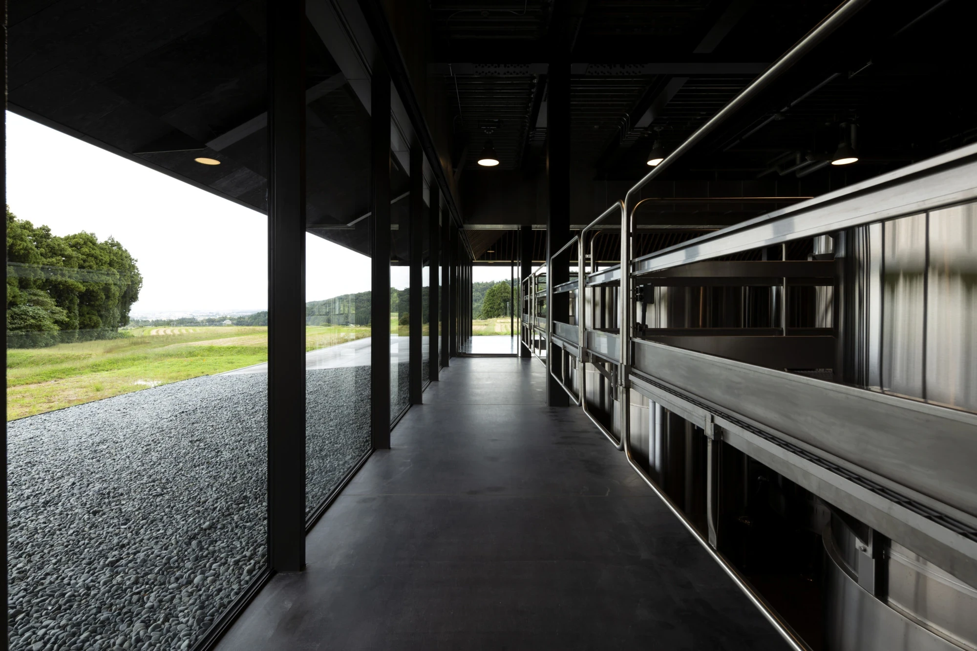 IWA Sake Brewery by Kengo Kuma & Associates, photographed by Mitsumasa Fujitsuka — A dark glazed circulation gallery runs the length of the tank hall, steel vessels ranked along one side and the rice fields pressed against the glass on the other