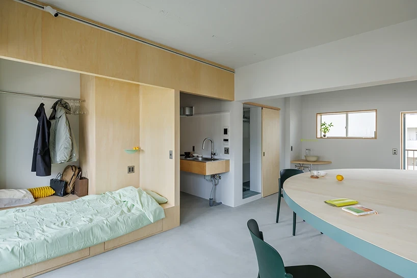 A Large Semicircle That Holds Everyday Life by Tato Architects, photographed by Yohei Sasakura: the plywood bedroom volume with sliding doors, wardrobe and mint bedding sits at left, kitchen behind, the table's curved edge and teal at right.