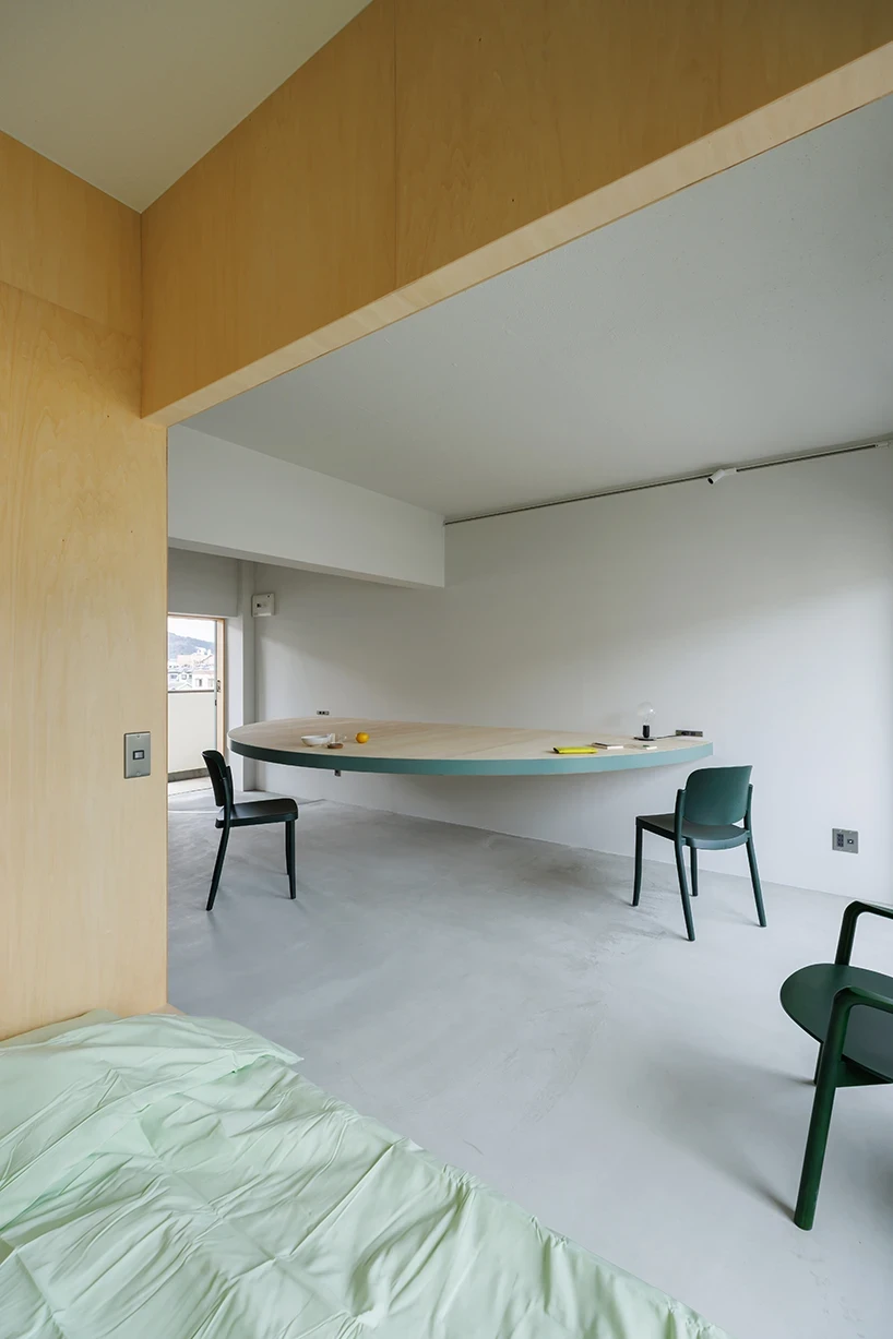 A Large Semicircle That Holds Everyday Life by Tato Architects, photographed by Yohei Sasakura: seen through the plywood opening from the bedroom side, the floating table with three teal chairs beyond the mint bed in the foreground.